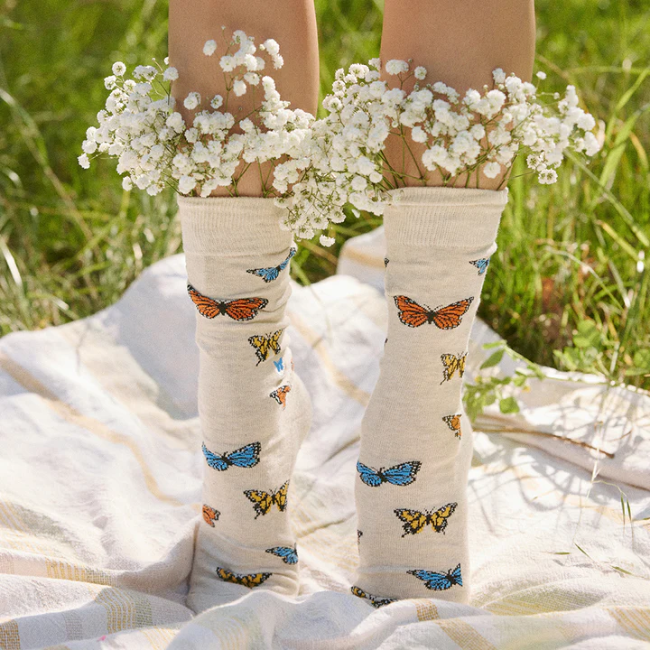 A person is shown wearing light-colored socks with a multi-colored butterfly pattern. Bunches of white flowers are tucked into the top of the socks. The person is standing on a checkered blanket in a grassy area.