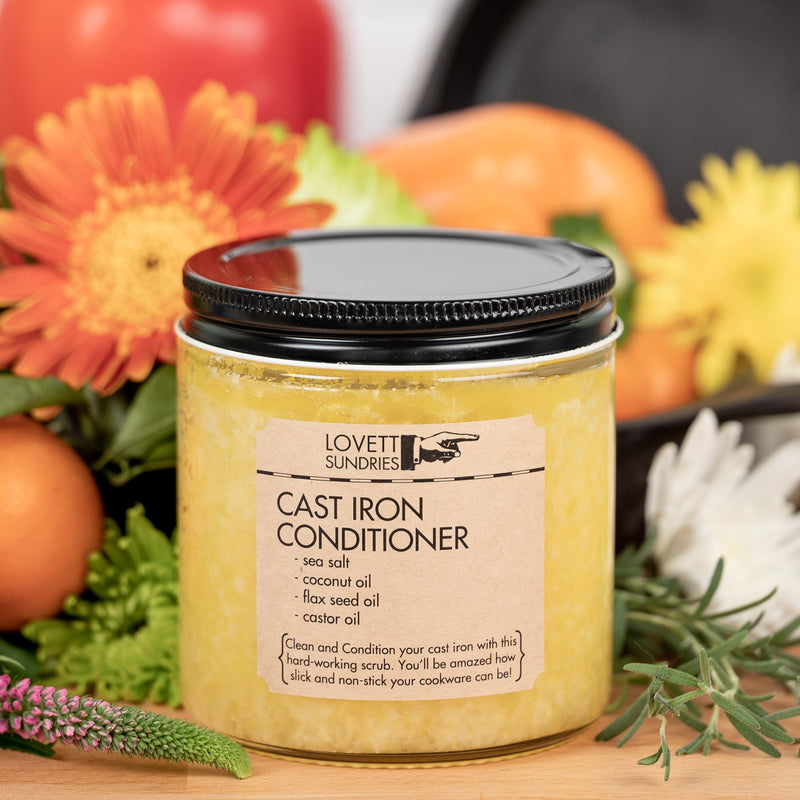 A jar of "Lovett Sundries Cast Iron Conditioner" with a black lid rests on a bamboo counter. It's surrounded by fresh flowers and vegetables to create a kitchen scene.