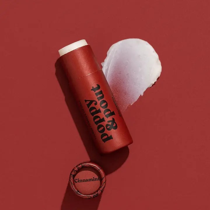 Poppy & Pout Cinnamint lip balm on a carmine surface, featuring its warm cinnamon-colored tube and vintage-inspired font label.