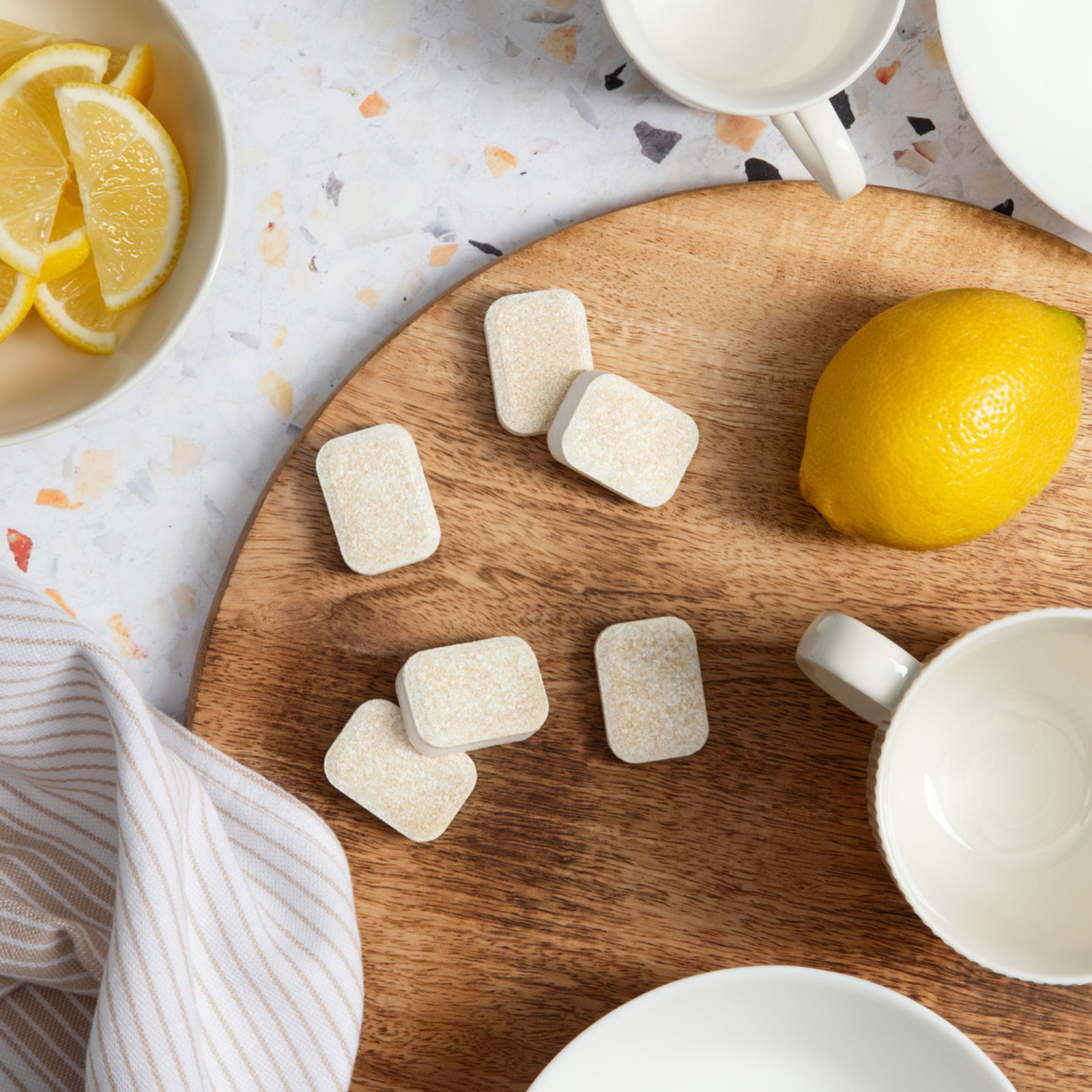 A collection of textured, light-colored dishwasher tablets are scattered across a round wooden cutting board, hinting at a natural and effective cleaning solution. A fresh lemon, a bowl of sliced lemons and a couple of white ceramic mugs are arranged nearby, suggesting a clean, citrus-fresh result and a simple, appealing aesthetic.