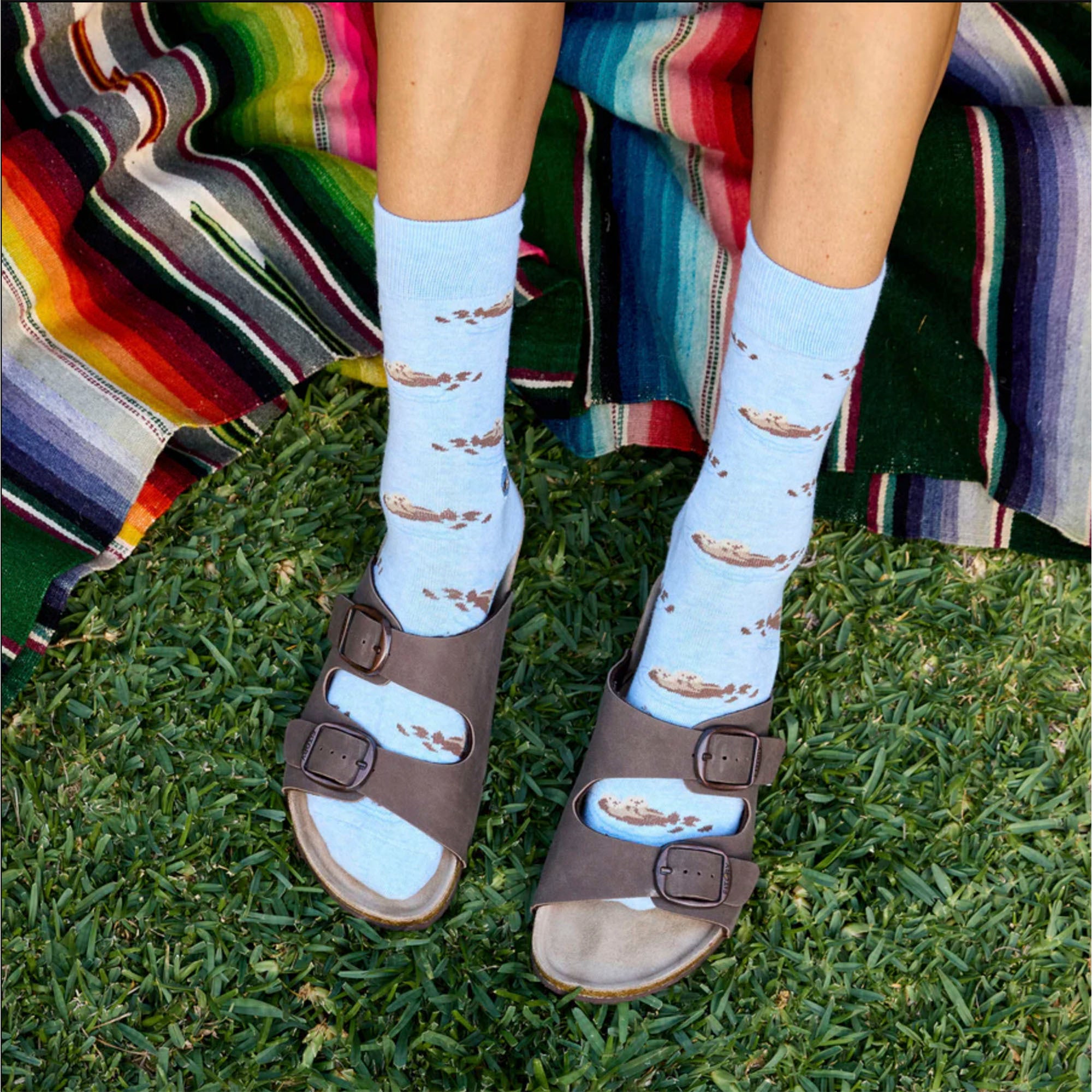 A person is shown wearing light blue socks with a sea otter pattern and brown sandals. The person is seated on green grass with a colorful striped blanket in the background.
