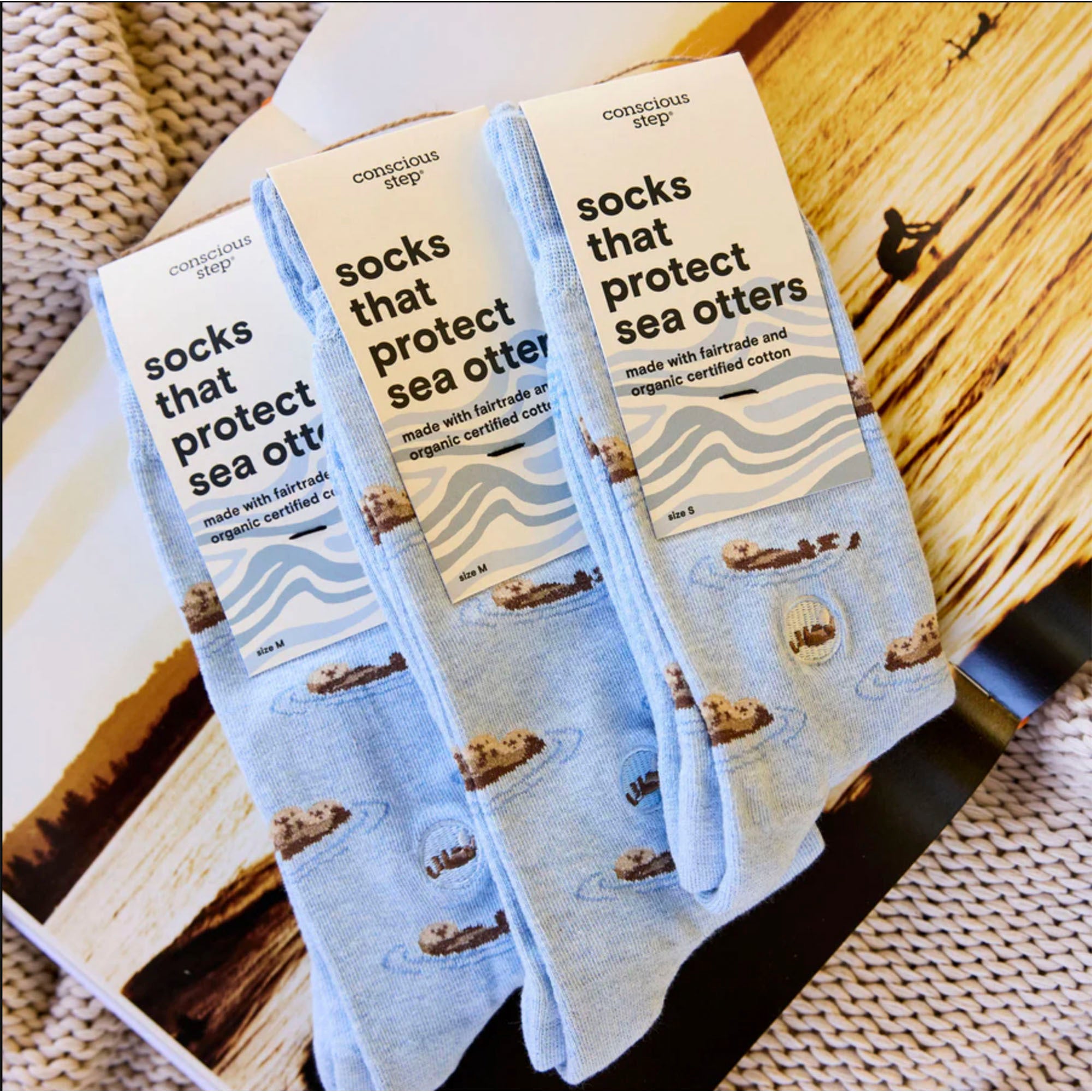 The image shows three pairs of light blue Conscious Step socks displayed on a surface. The socks are patterned with sea otters floating on their backs and have tags that read "socks that protect sea otters". The tags also state that the socks are made with fairtrade and organic certified cotton, with sizes listed as M and S.