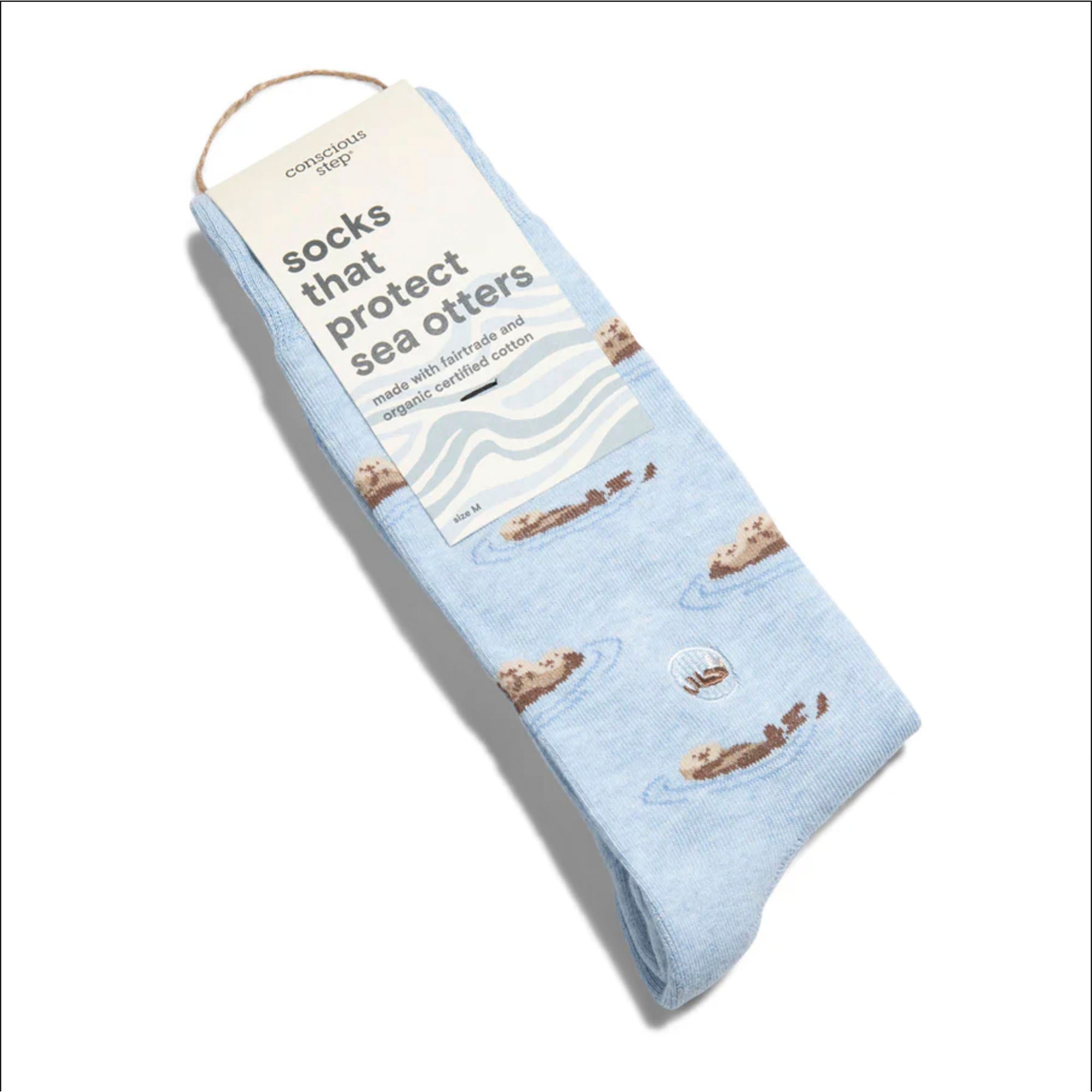 A pair of light blue Conscious Step socks with a sea otter pattern is shown with a tag. The tag identifies them as "socks that protect sea otters" and notes they are made with fairtrade and organic certified cotton.