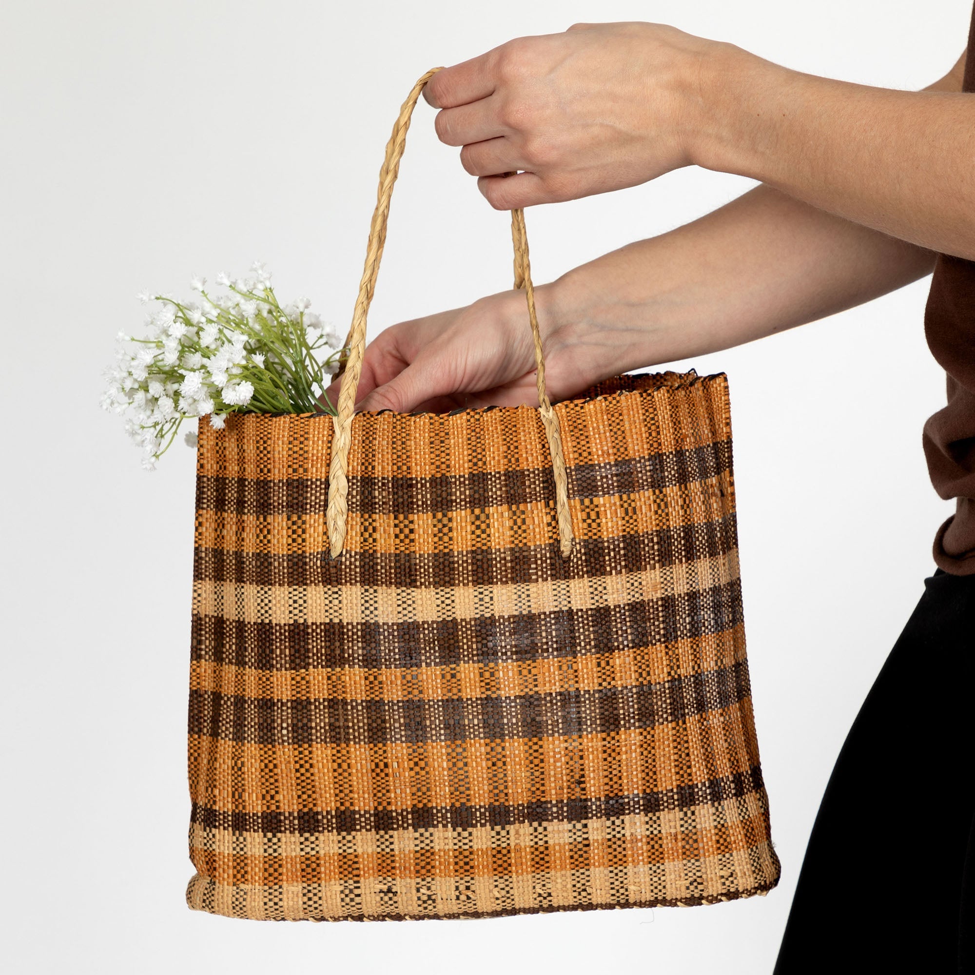 Person holding a natural grass tote bag showcasing its earthy hues and braided handles. Handmade by Congolese artisans using vegetable-dyed fibers for a lightweight and durable carry-all.