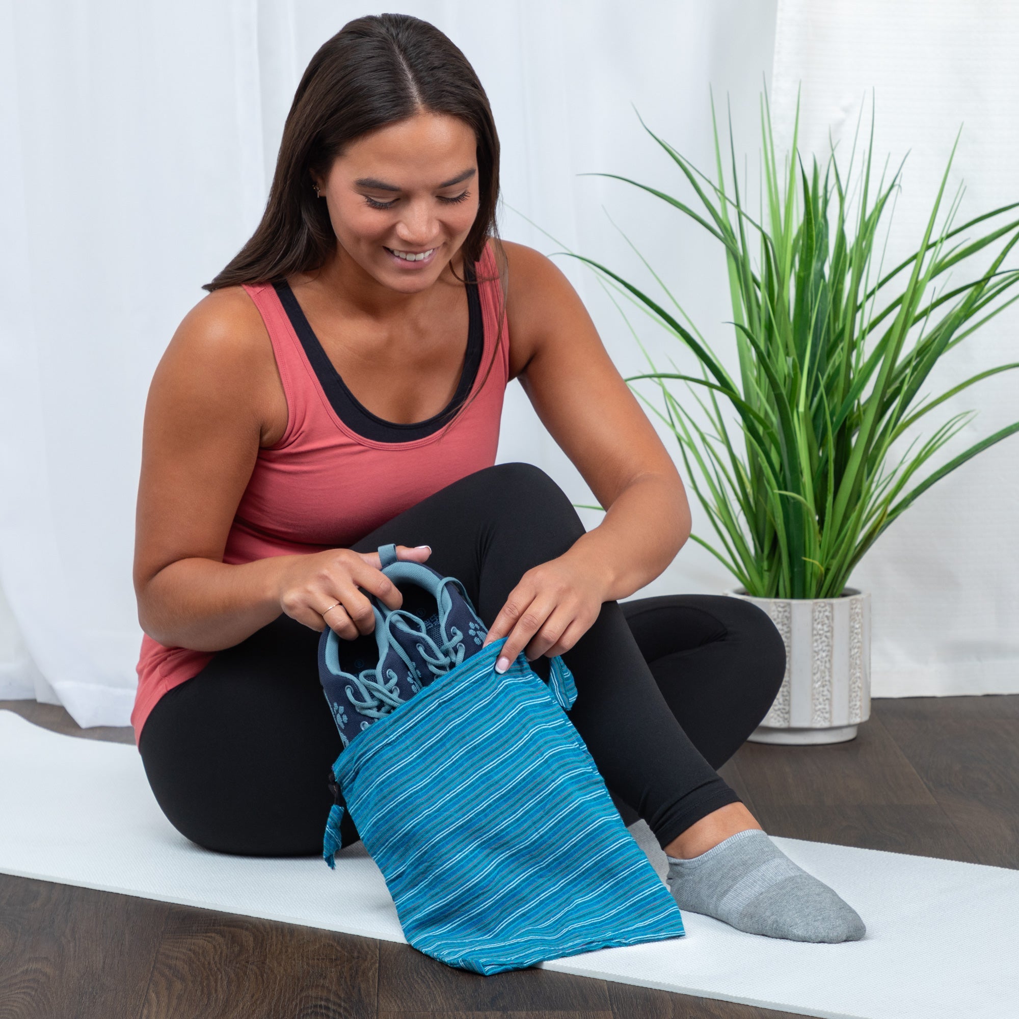 Person packing a pair of white sneakers into a recycled drawstring Shoe Bag on a wooden bench, with a potted plant and natural light in the background.