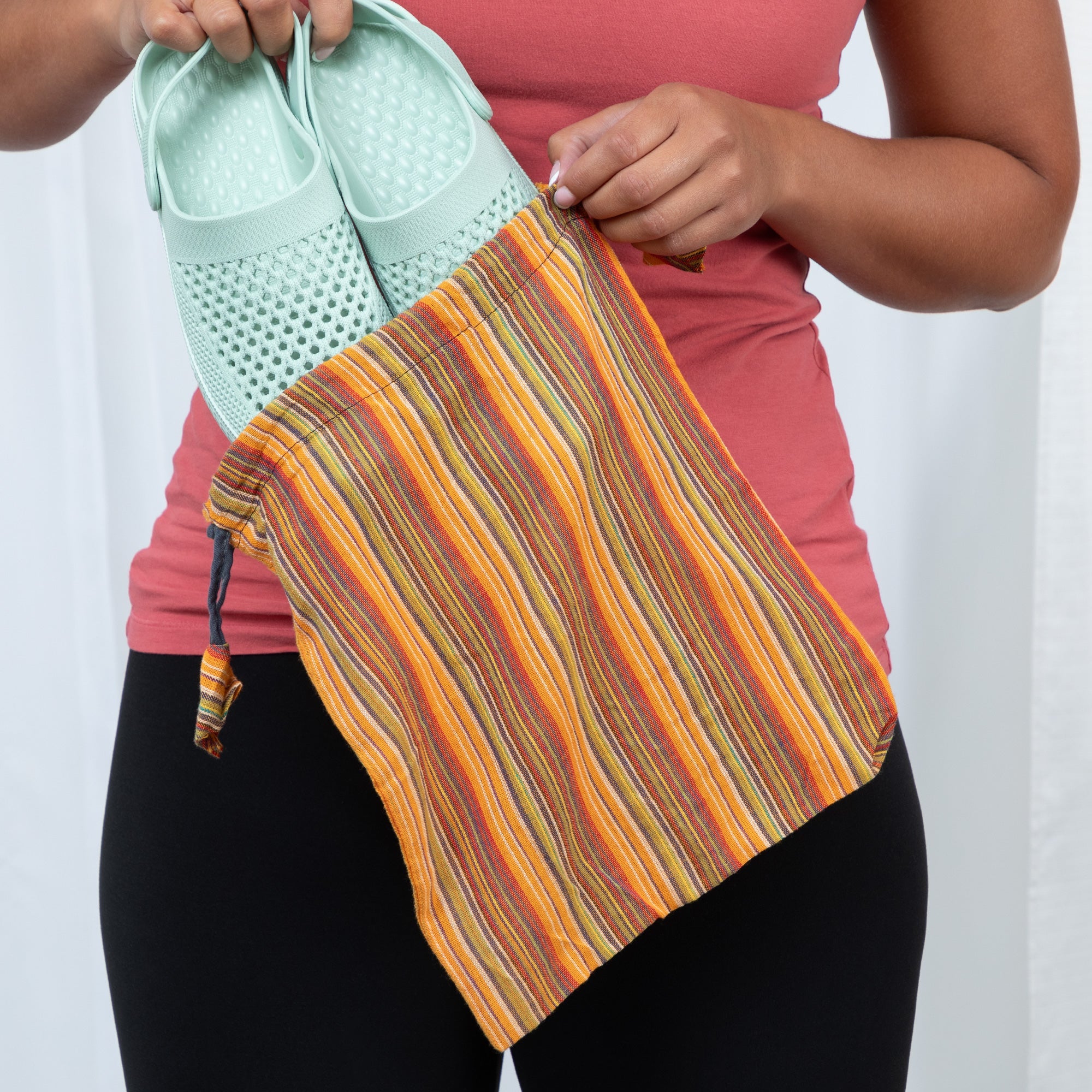A person places teal slip-on shoes into a yellow and orange striped recycled drawstring bag, showing its utility for eco-friendly organization.