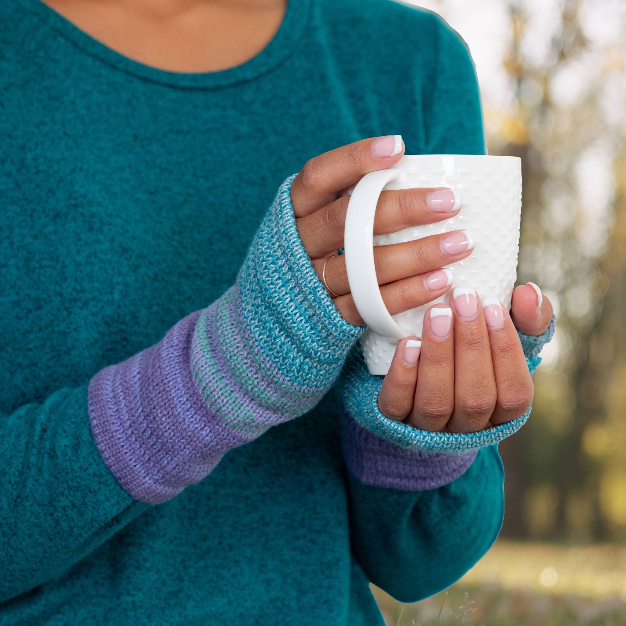 Hands holding a cup of coffee while wearing alpaca fingerless mittens in blue, gray, and purple. Lightweight and naturally insulating.