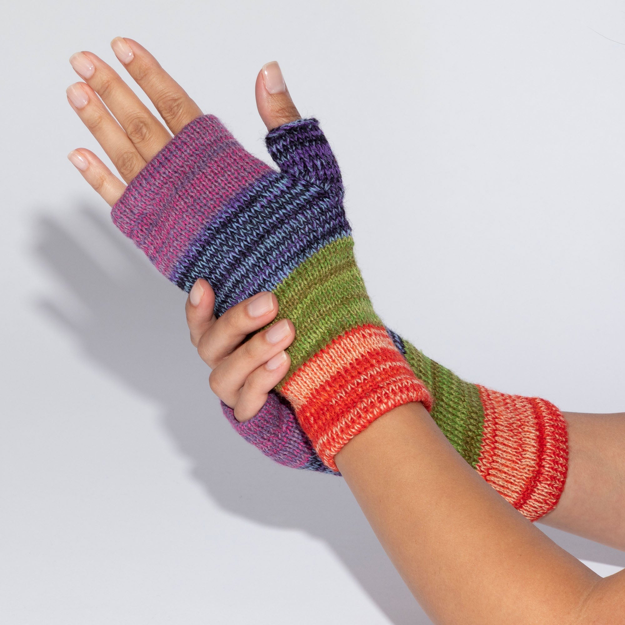 Cose-up of hands wearing handmade alpaca fingerless mittens with a striped knit pattern in purple, blue, green, and red.
