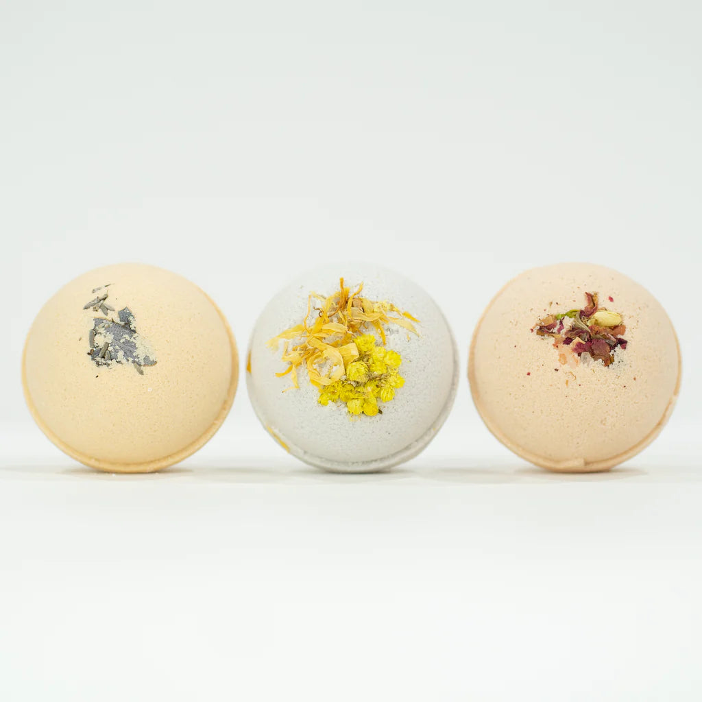 Three botanical bath bombs are shown in a line against a white background. Each bath bomb is topped with a different arrangement of dried flowers and herbs.