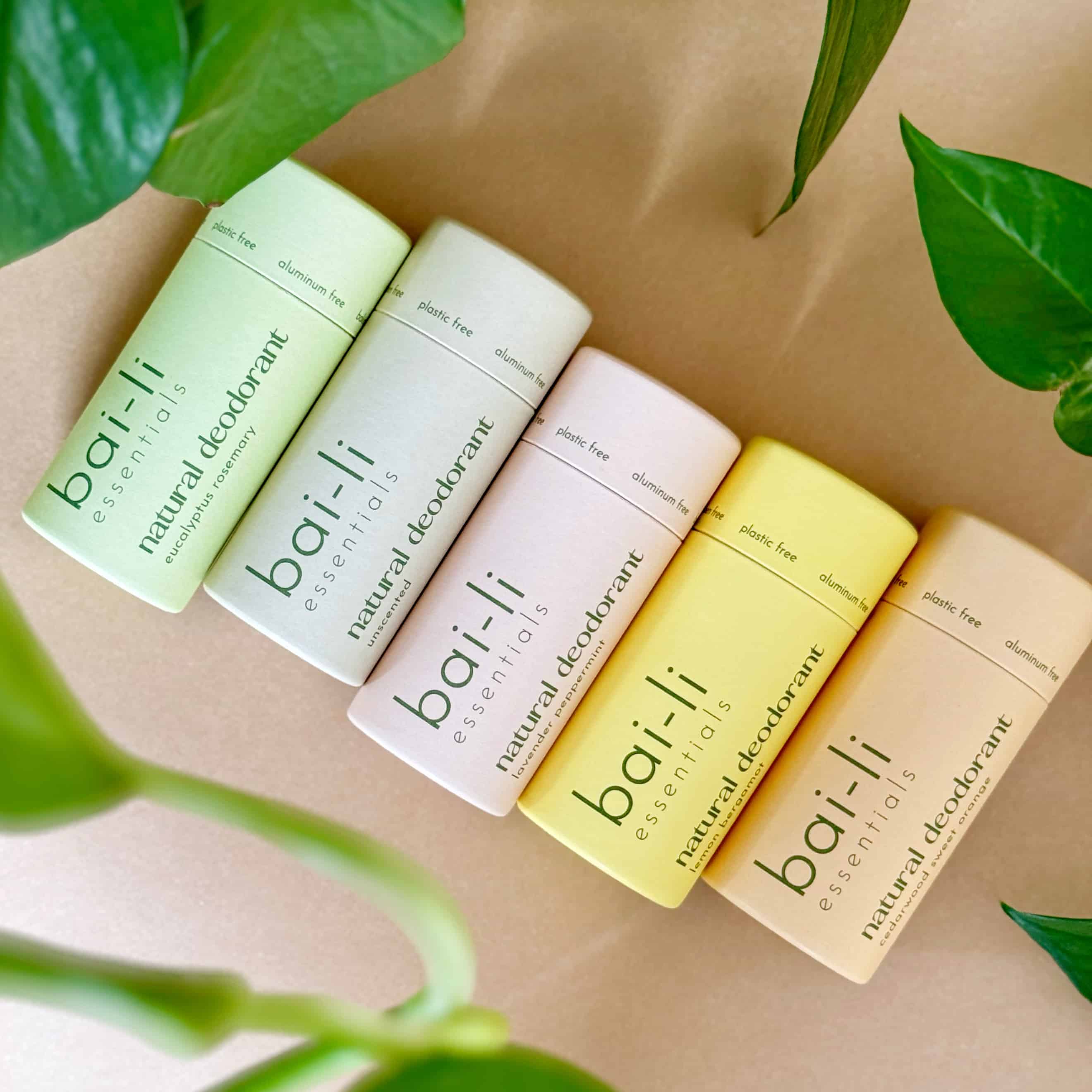 Top-down view of five plastic-free deodorant tubes arranged on a light surface surrounded by green plants. Scents included are Lemon + Bergamot, Lavender + Peppermint, Cedarwood + Sweet Orange, Eucalyptus + Rosemary, and Unscented. Made with organic ingredients and compostable packaging.