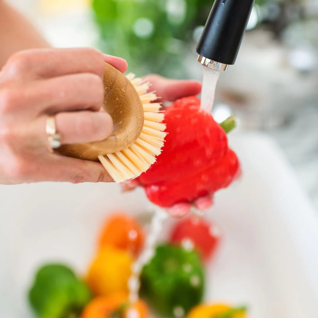 A close up shot of a hand holding a bamboo vegetable brush with light-colored bristles scrubbing a bright red bell pepper under running water from a matte black faucet. Other colorful peppers are blurred in the background of the white sink.