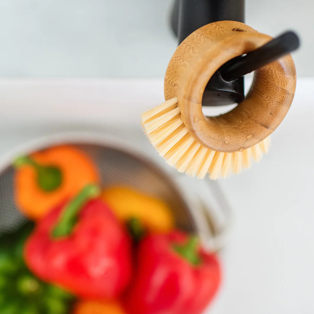 A bamboo veggie brush is hanging on a matte black faucet above a kitchen sink. The bristles are a light blonde color. Below in the sink a mesh strainer is filled with red orange and yellow bell peppers ready to be cleaned.