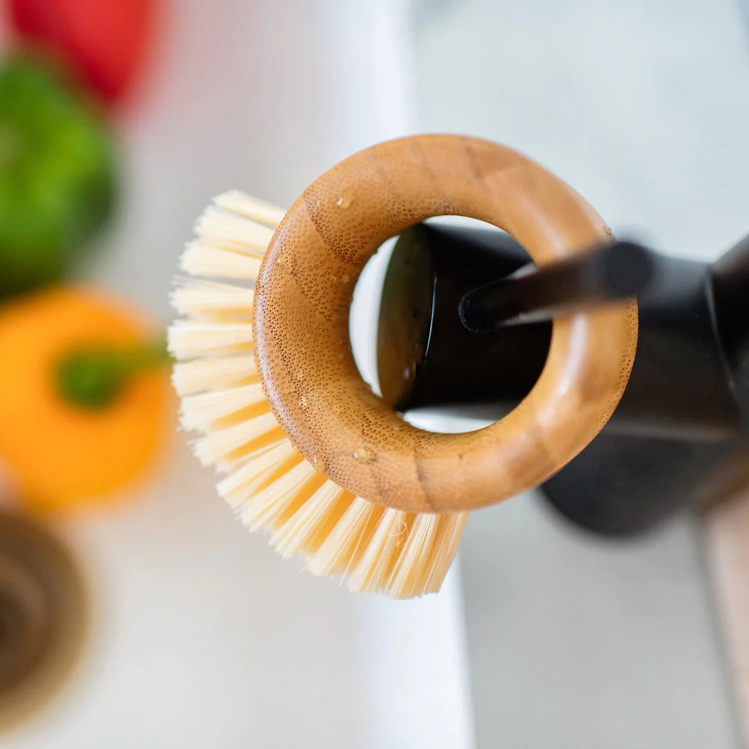 A circular bamboo vegetable brush with light-colored bristles is hanging from a matte black faucet over a white sink. There are small droplets of water on the bamboo. The blurred background shows vegetables in the sink.