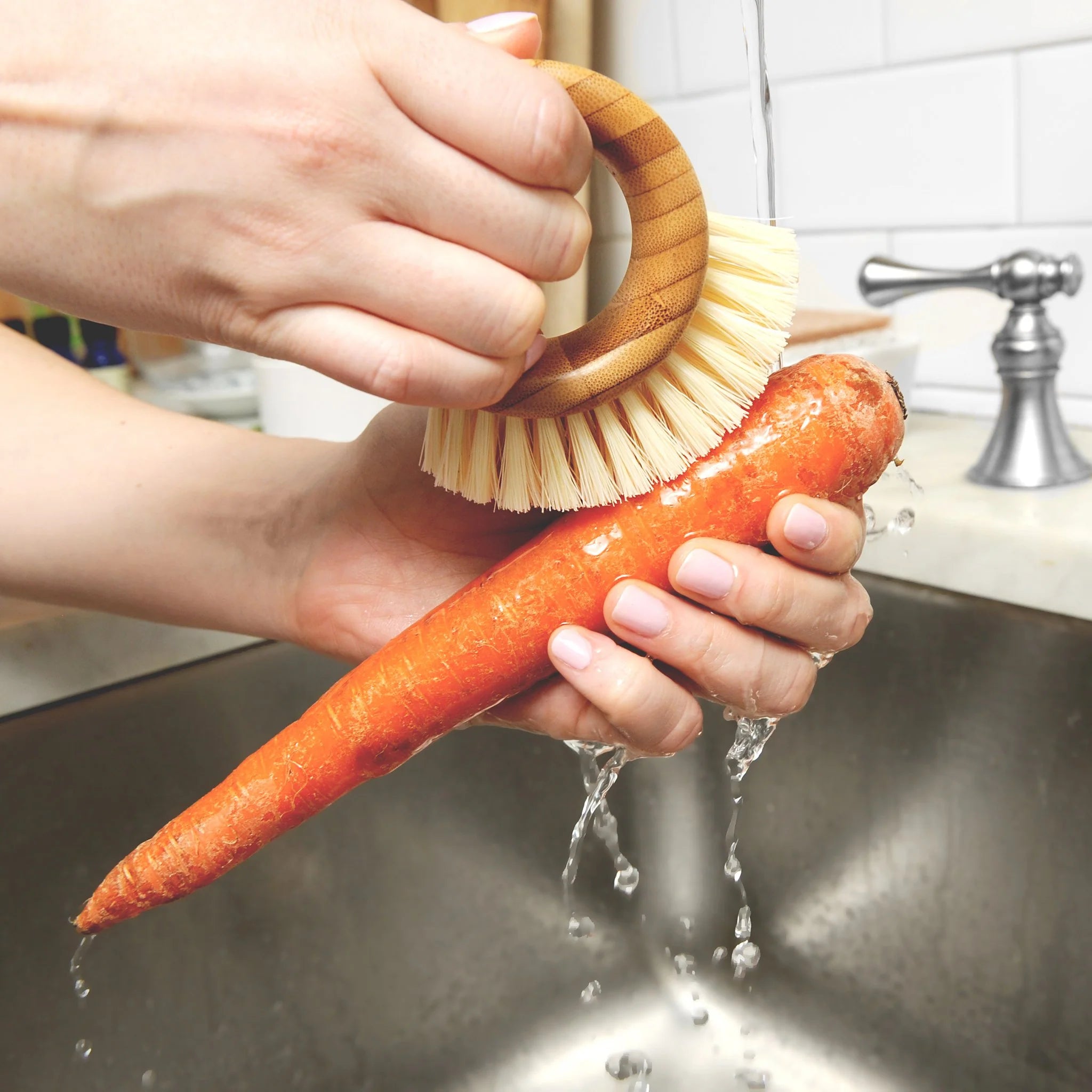 A pair of hands holds a large orange carrot under a stream of water from a faucet in a stainless steel sink. The right hand grips a ring-shaped bamboo veggie brush with blonde bristles to scrub the carrot clean.