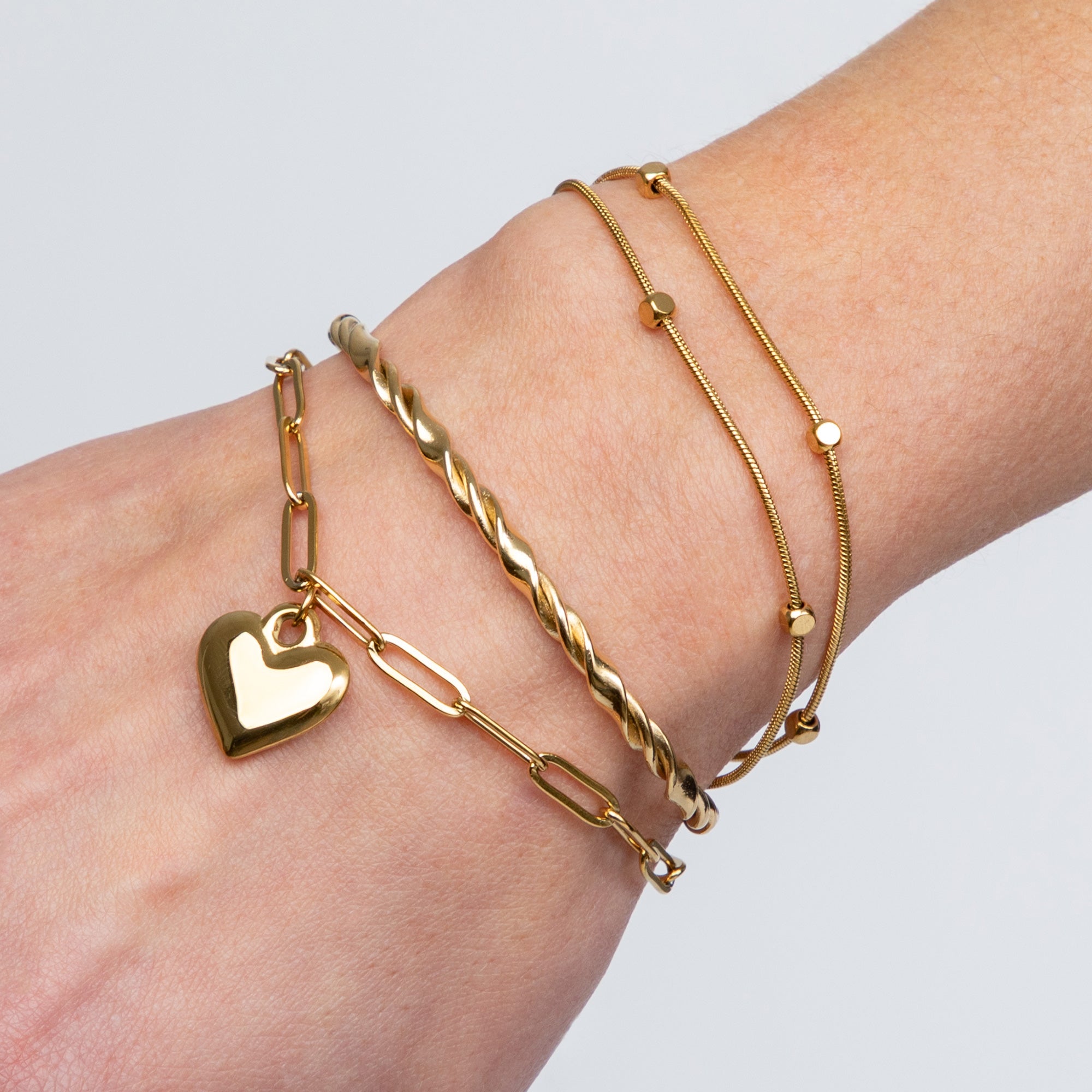 A hand wearing four recycled 14K gold and stainless steel bracelets—Shell Ya!, Twisted Gold Cuff, Ocean Love with a heart charm, and Double Strand Cube—each showcasing elegant, eco-friendly craftsmanship and water-resistant shine.