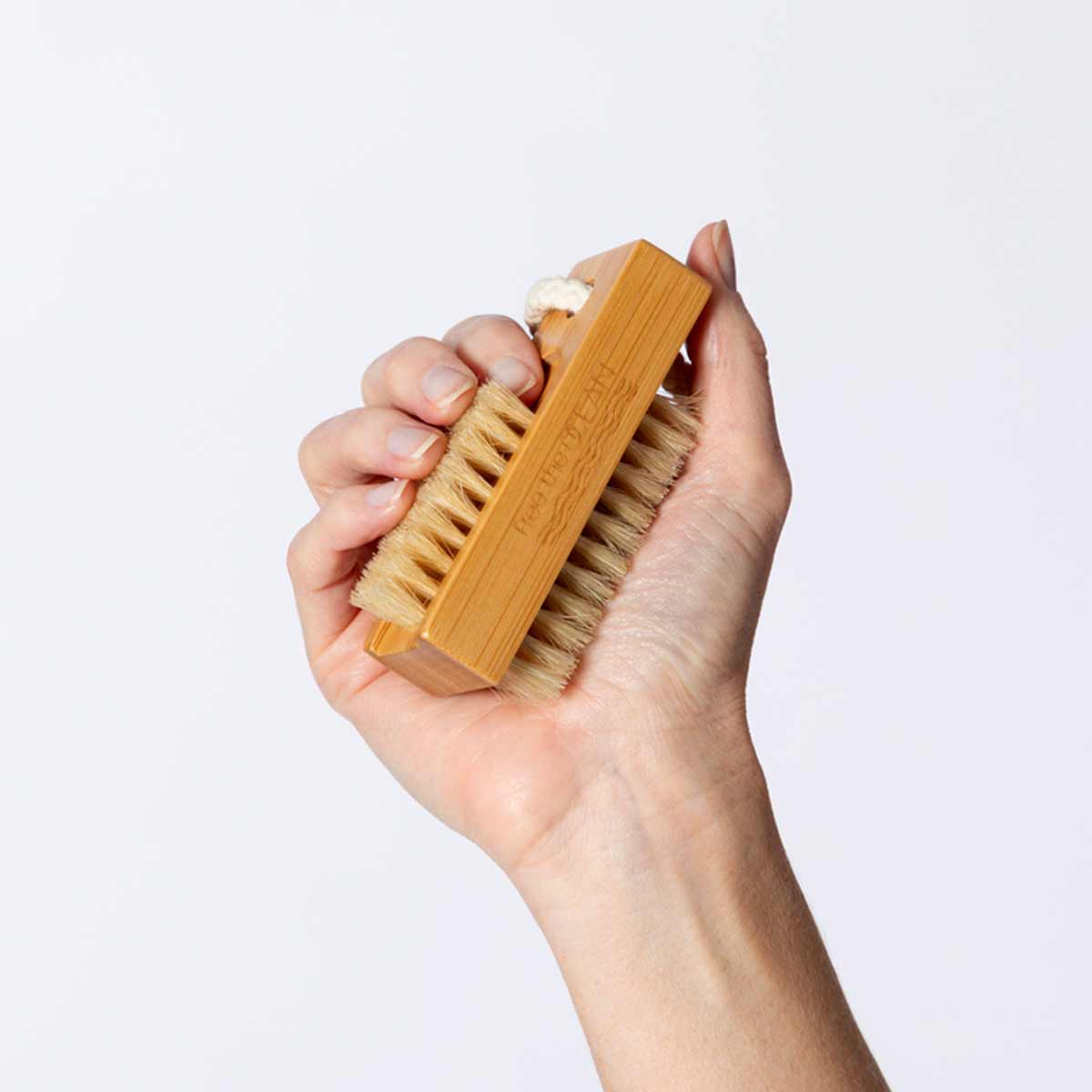 A hand is holding a bamboo brush. The brush has two sets of bristles and a braided rope loop attached to one end, which is visible at the top. The image helps to show the size of the brush relative to a hand.