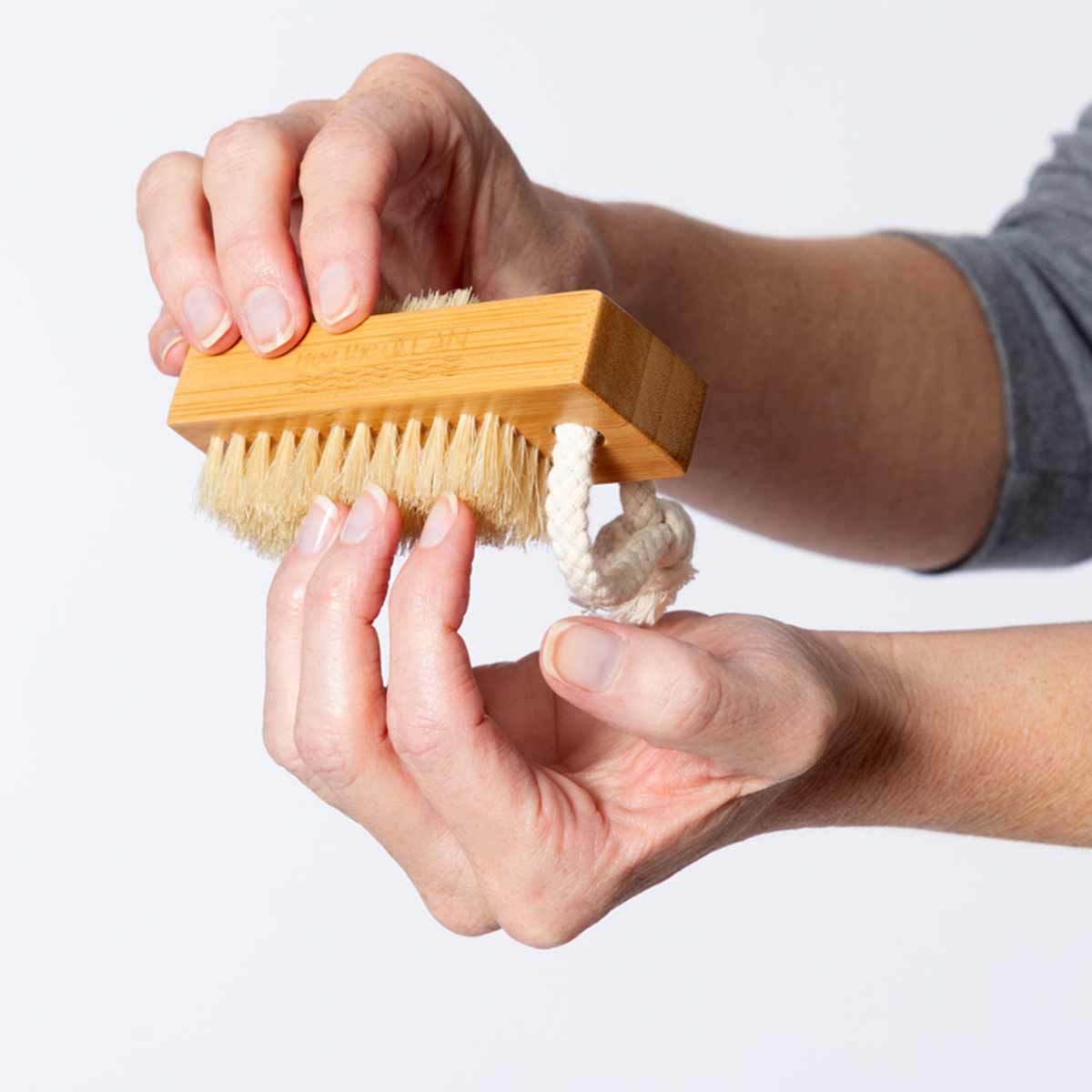 Two hands are holding and demonstrating the use of a bamboo nail brush with a braided rope loop. The brush has two sets of bristles and an etched logo on its handle.