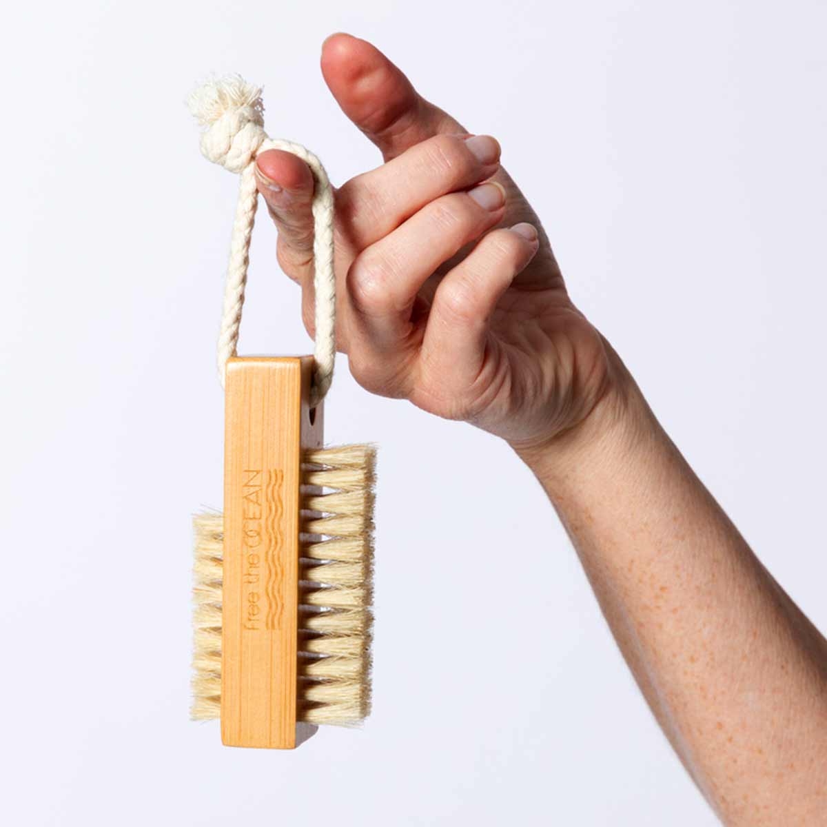 A hand is holding a bamboo brush with a braided rope loop attached to it. The Free the OCEAN logo is visible on the side of the handle. This image shows that the brush is almost the size of the hand holding it.
