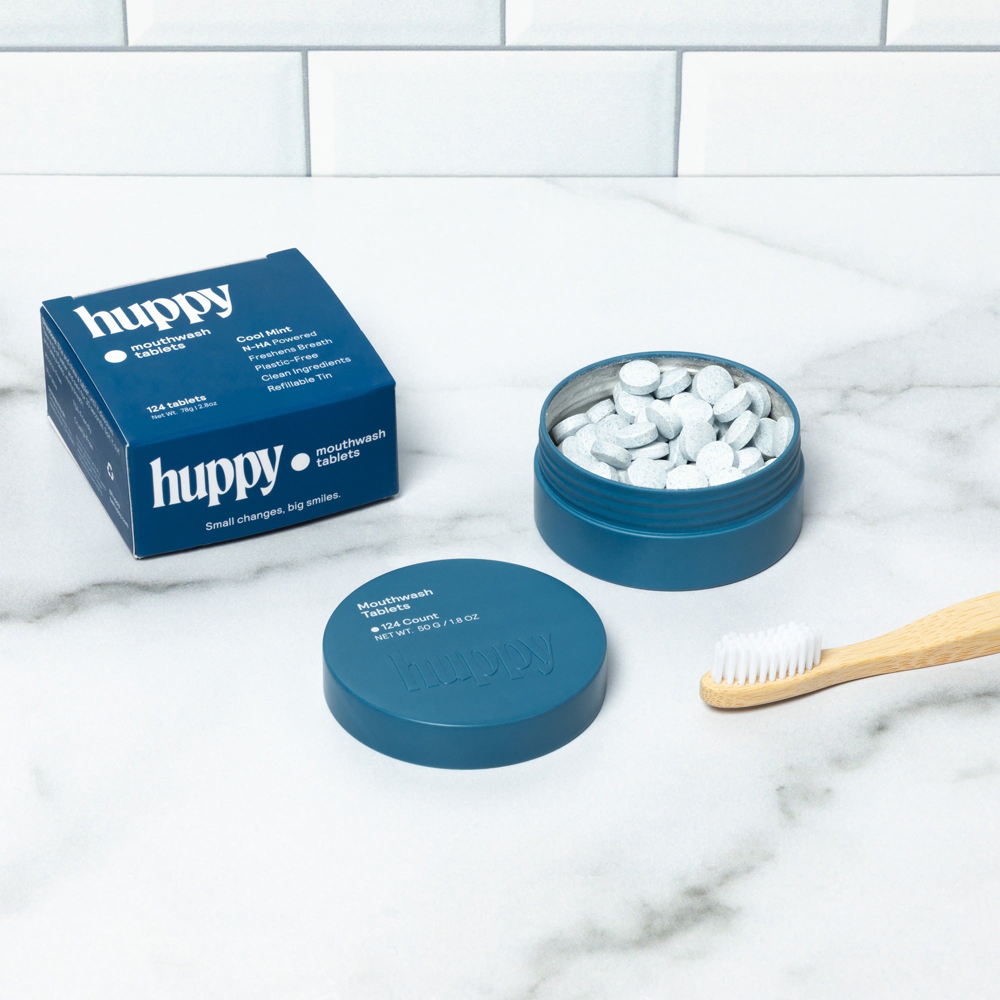A cute flat-lay of Huppy mouthwash products. A blue tin of tablets is open next to its lid and a bamboo toothbrush. A blue refill box is also present, completing the eco-friendly dental set.
