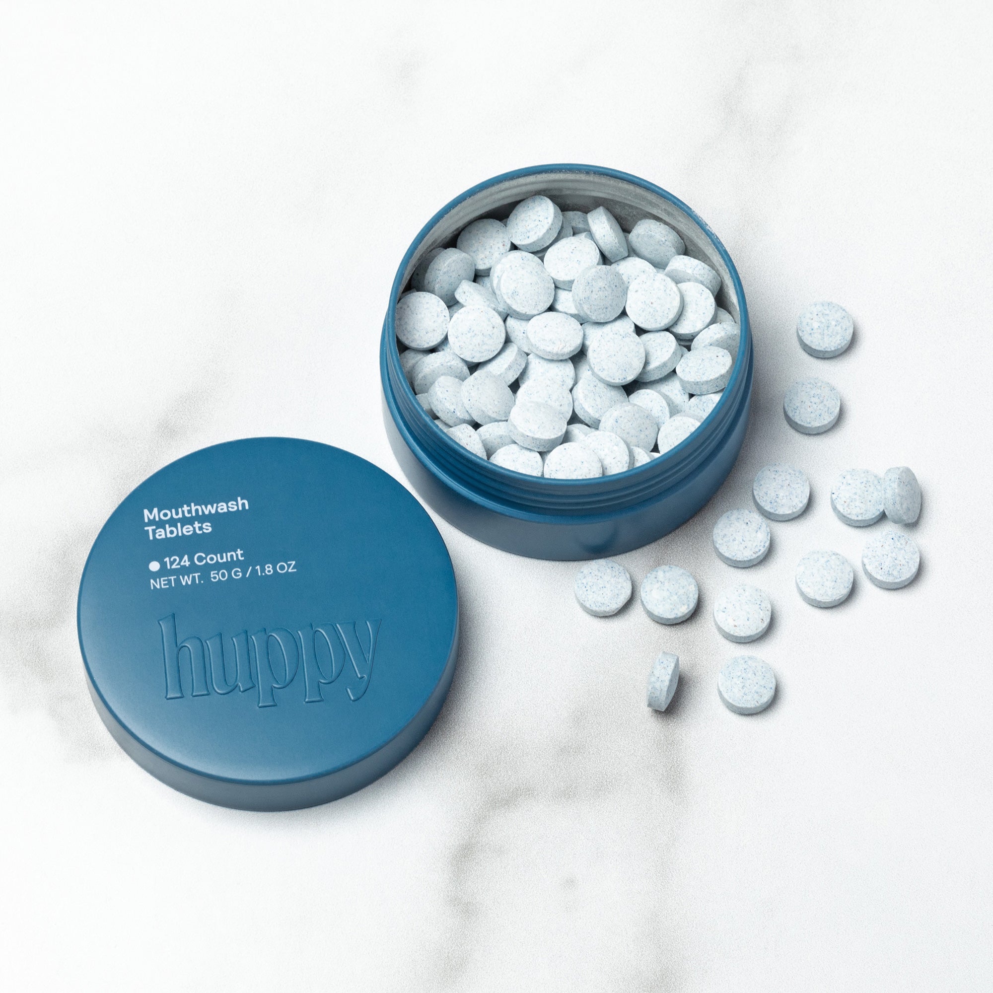 A cute blue tin of Huppy Mouthwash Tablets is open, showing small, speckled blue-gray tablets inside and scattered around the tin. The lid, which has the text "124 Count" is next to it.