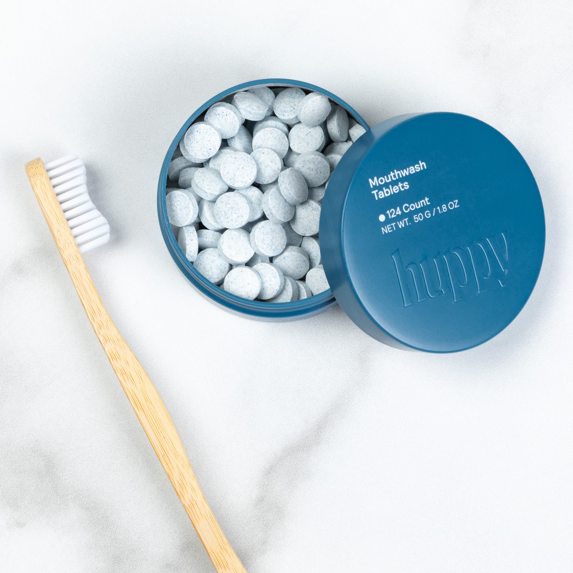 A cute blue tin of Huppy Mouthwash Tablets is open next to its lid and a bamboo toothbrush. The tin is full of speckled blue-gray tablets, and the lid reads "124 Count".