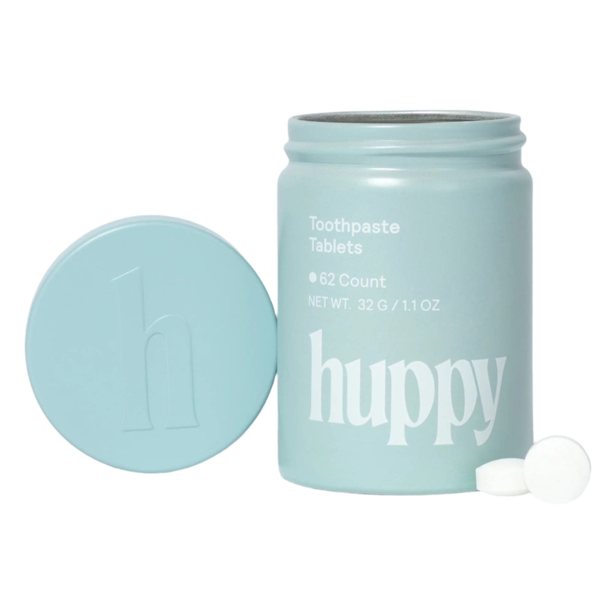 A light blue 'huppy' jar of toothpaste tablets has its lid off, with two small white tablets sitting beside it.