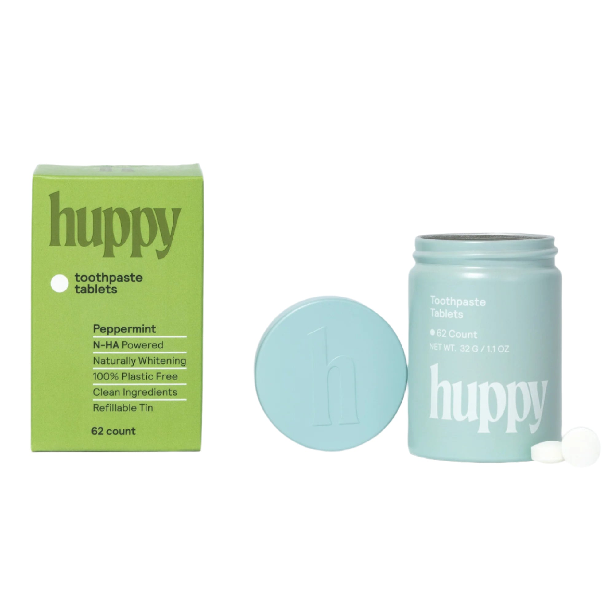 A green cardboard box of 'huppy' peppermint toothpaste tablets stands next to a small, light blue refillable tin. The box mentions "N-HA Powered" and "100% Plastic Free," with 62 tablets inside. Two round, white toothpaste tablets are next to the tin.