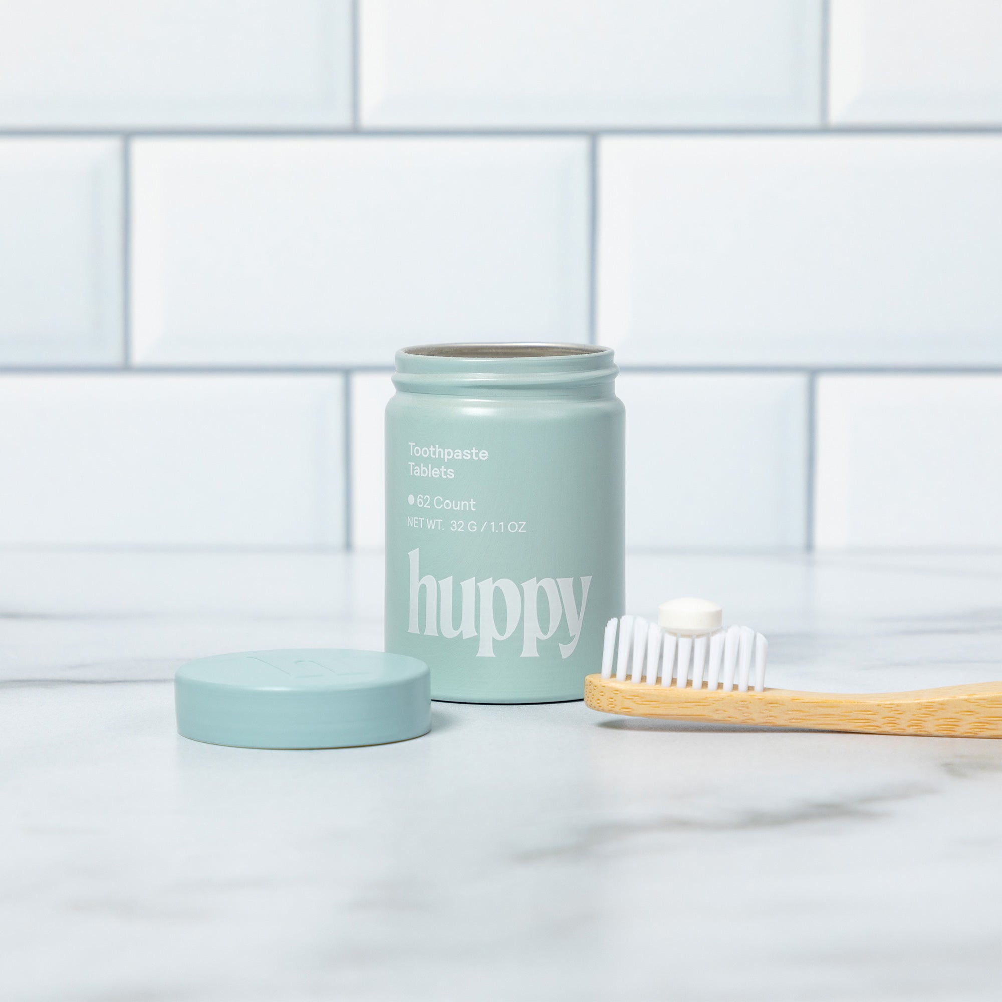 A 'huppy' toothpaste tablet jar with its lid off is on a counter next to a bamboo toothbrush with one tablet on its bristles.