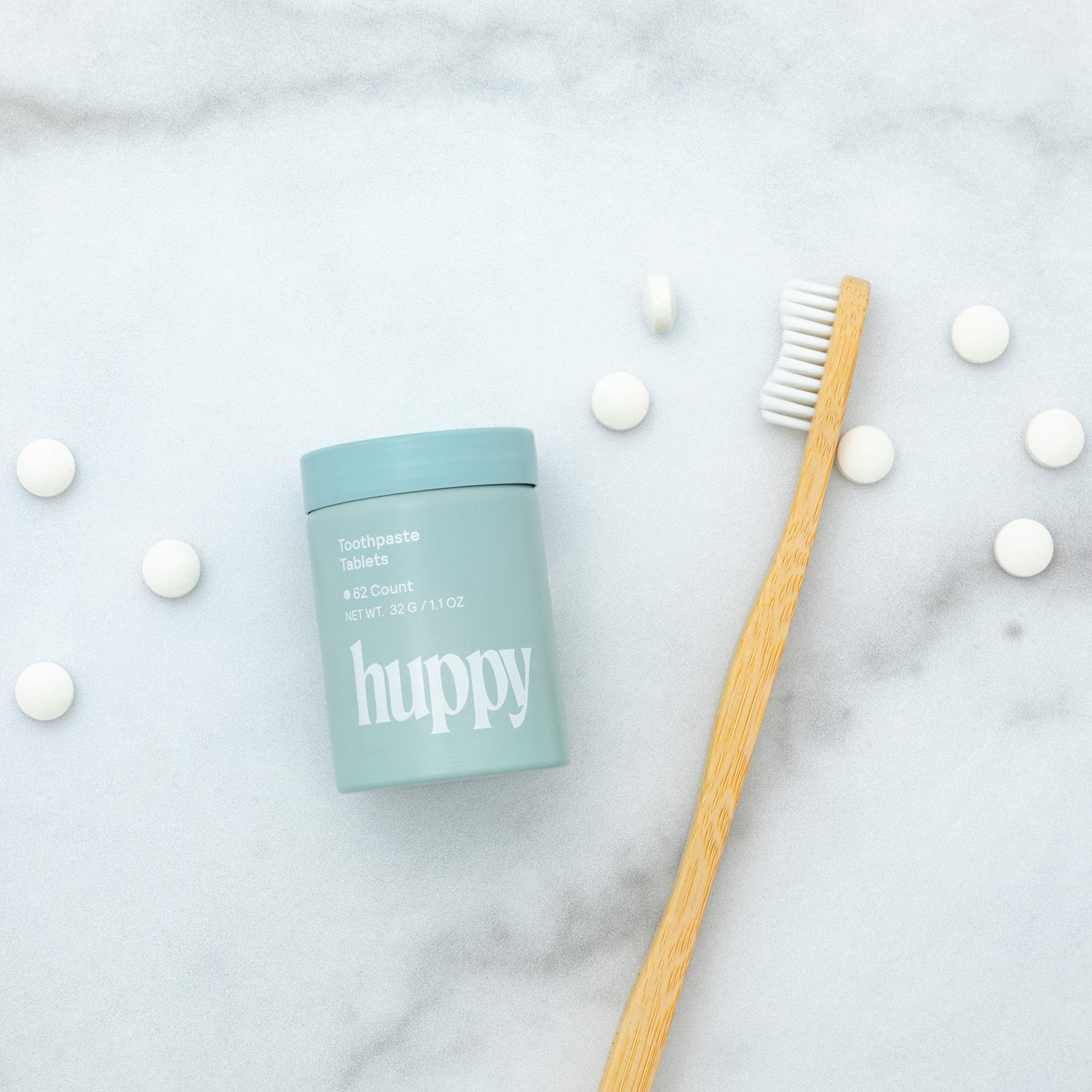 A light blue 'huppy' toothpaste tablet jar and a bamboo toothbrush are on a white marble surface, surrounded by many scattered white tablets.