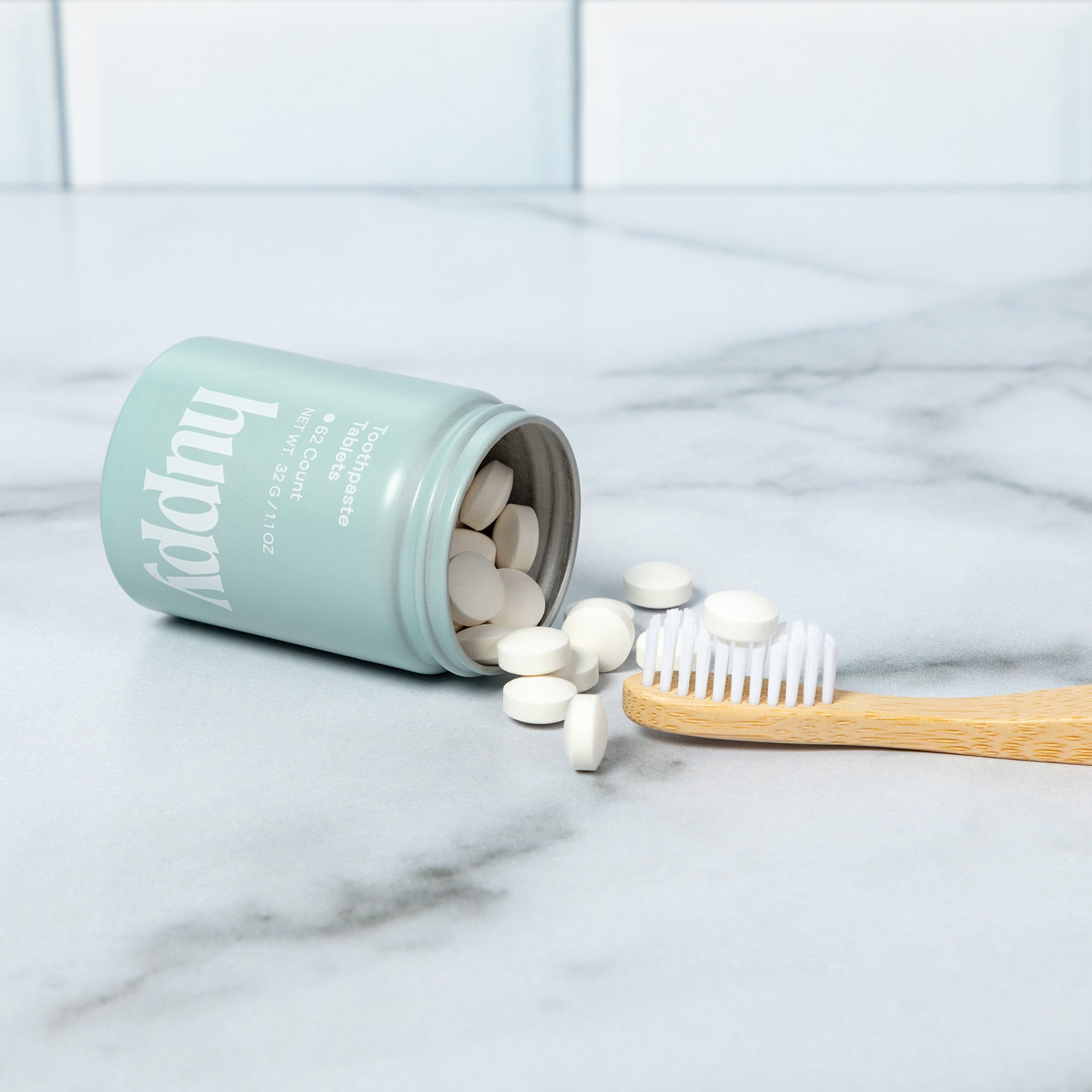 A 'huppy' jar lies on its side, spilling toothpaste tablets next to a bamboo toothbrush with one tablet on its bristles.