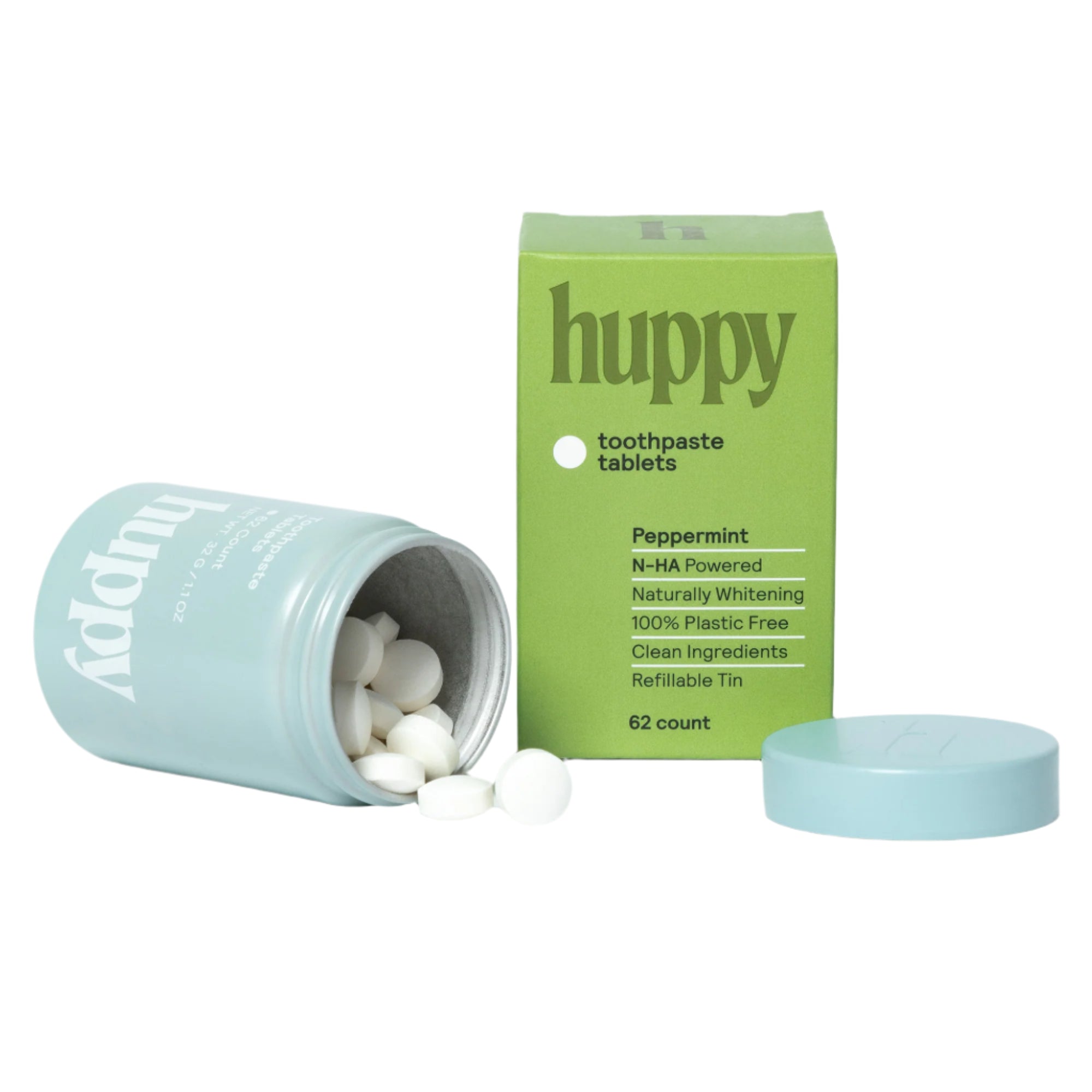A green 'huppy' toothpaste tablet box stands next to a tipped-over blue jar that is spilling its white tablets, with the lid beside them.
