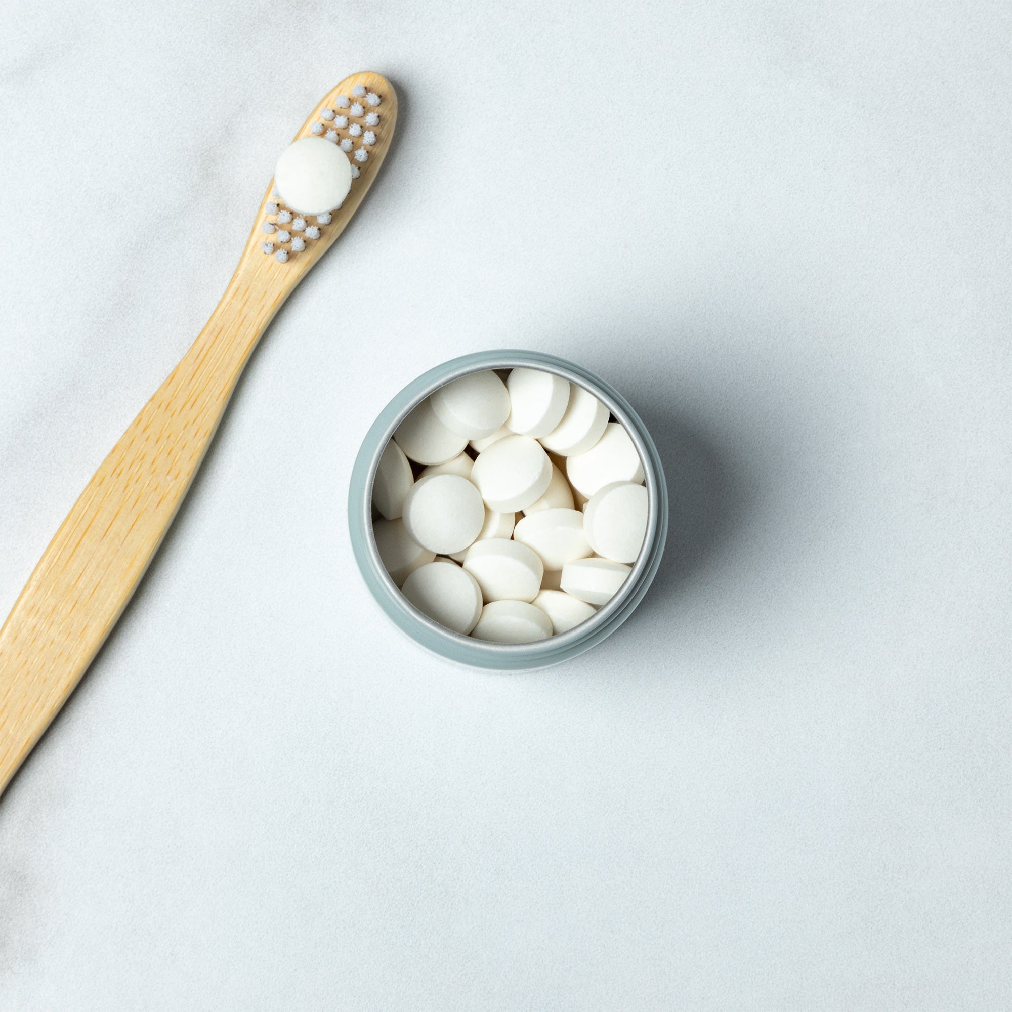 An open 'huppy' jar filled with white toothpaste tablets sits next to a bamboo toothbrush with one tablet on its bristles.