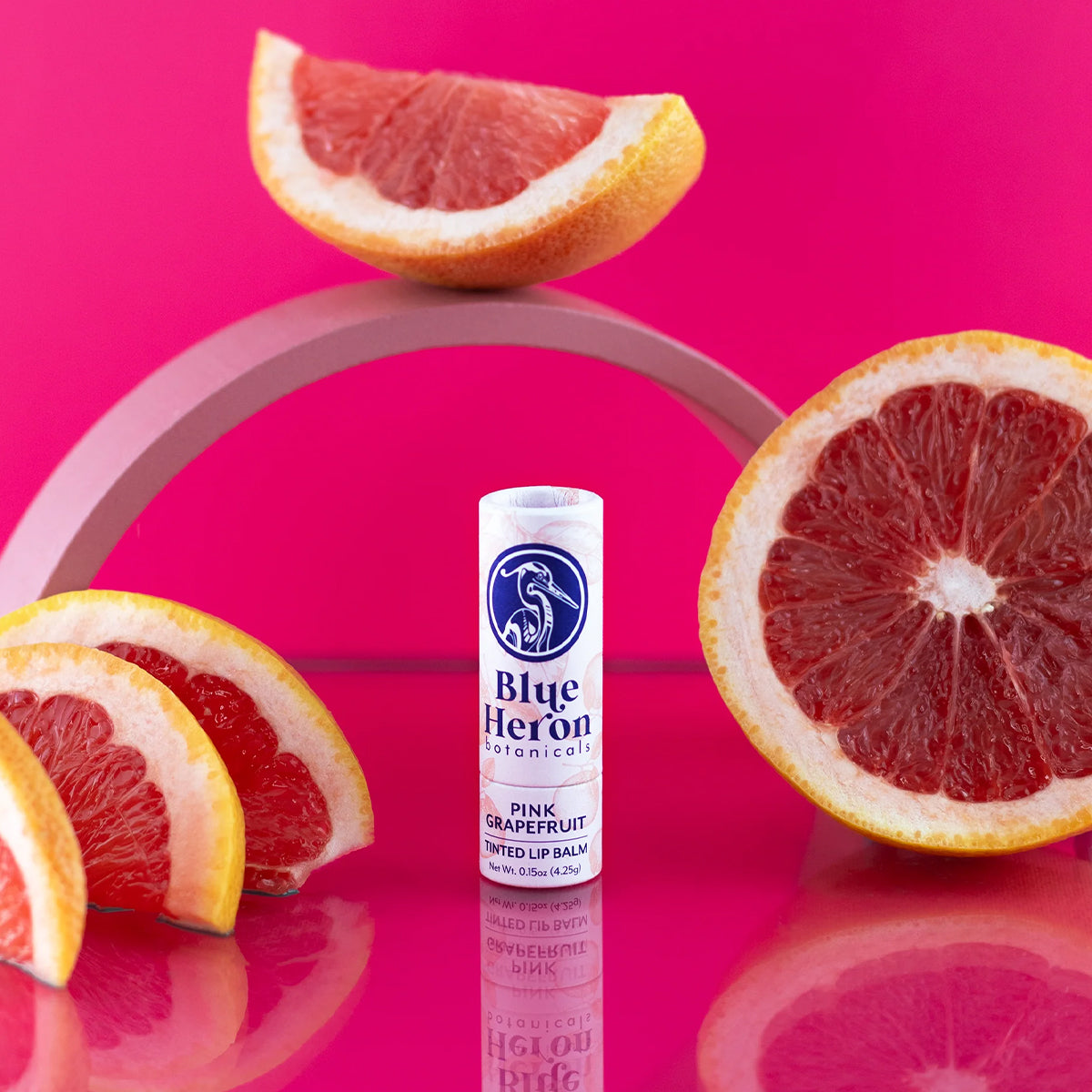 Pink grapefruit Blue Heron lip balm with pink grapefruit slices on a pink background