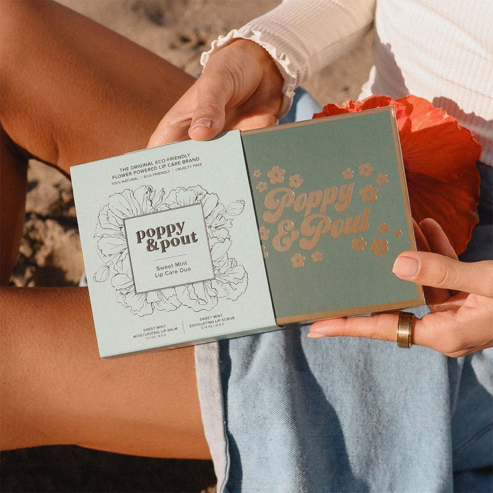 A lip balm package from 'Poppy & Pout' held by a person outdoors.