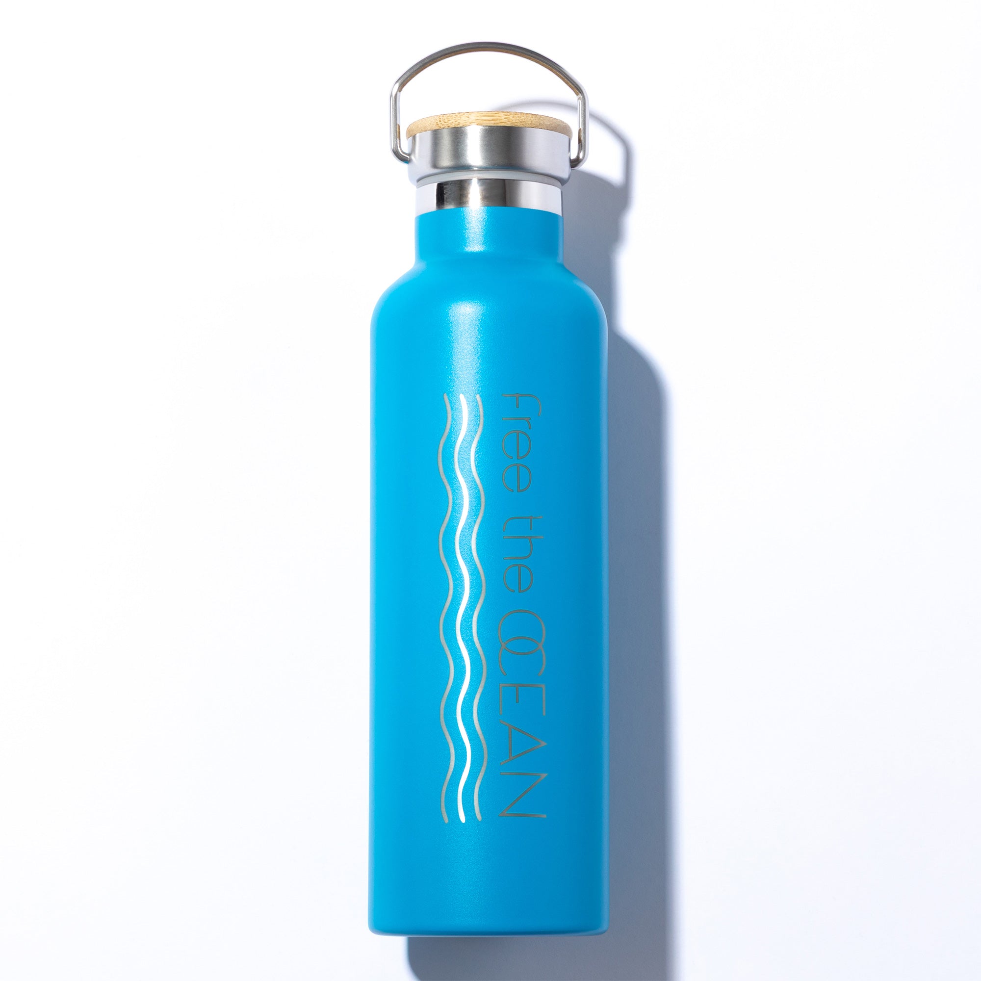 A turquoise reusable water bottle with a bamboo lid and a silver handle. It features two wavy silver lines and the phrase "Free the OCEAN" down the side.