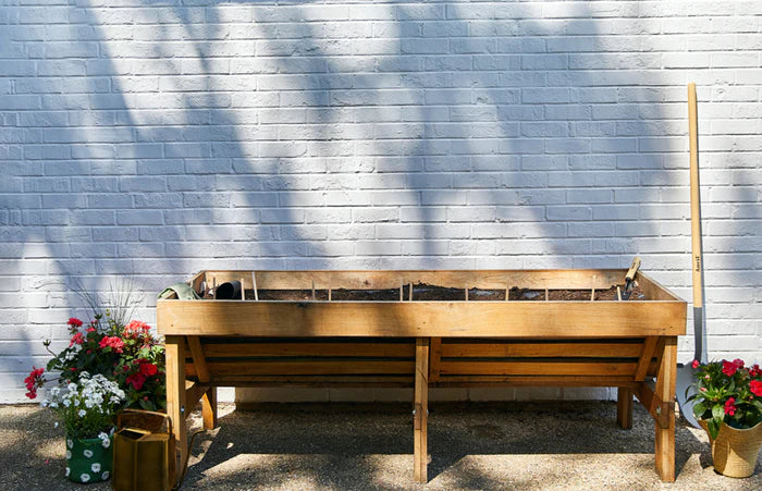 A clean, raised wooden garden bed filled with soil and garden seed pop sticks, ready for planting. It is set against a white brick wall, with potted plants and a gardening tool on either side.