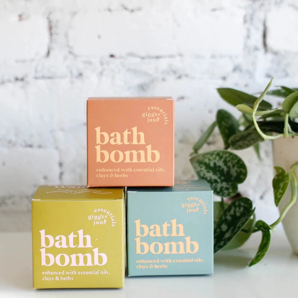 A pyramid stack of three Ginger June Essentials bath bomb boxes is shown in front of a white brick wall. The boxes are orange, light green and light blue, they are labeled "enhanced with essential oils, clays & herbs". A green plant is on the right side of the image.