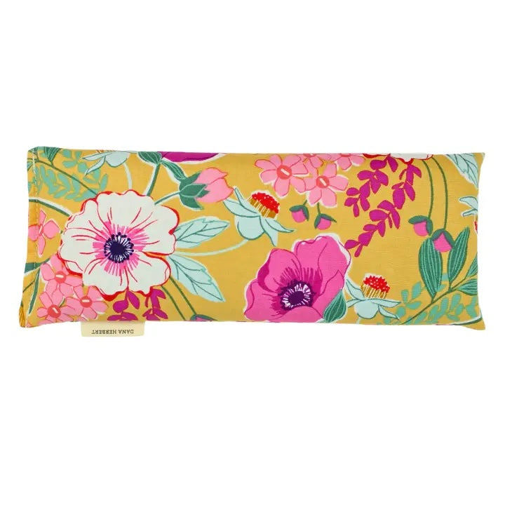 A rectangular eye pillow by Dana Herbert has a vibrant floral pattern with pink and white flowers on a mustard yellow background.