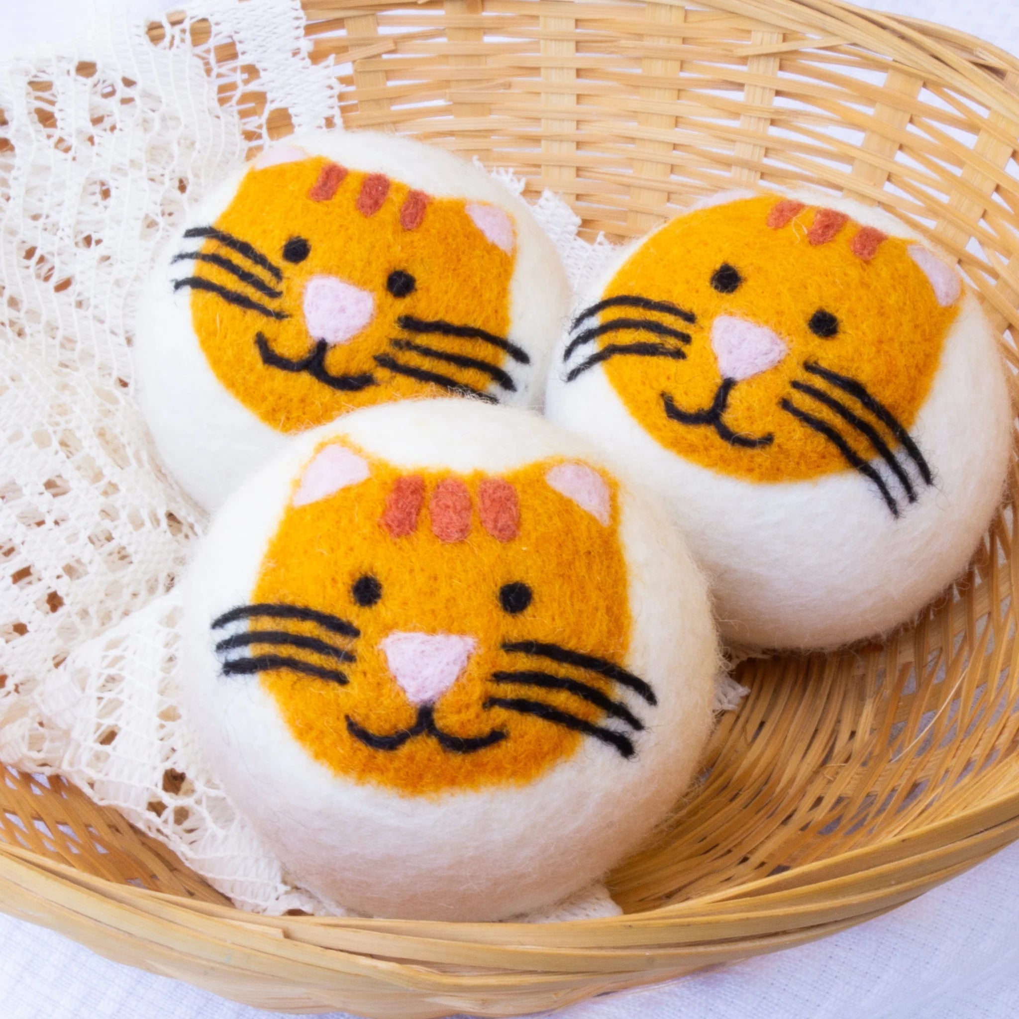A set of three adorable Friendsheep sustainable goods wool dryer balls, each decorated with a cute tiger face, are nestled together in a wicker basket on a white lace cloth.