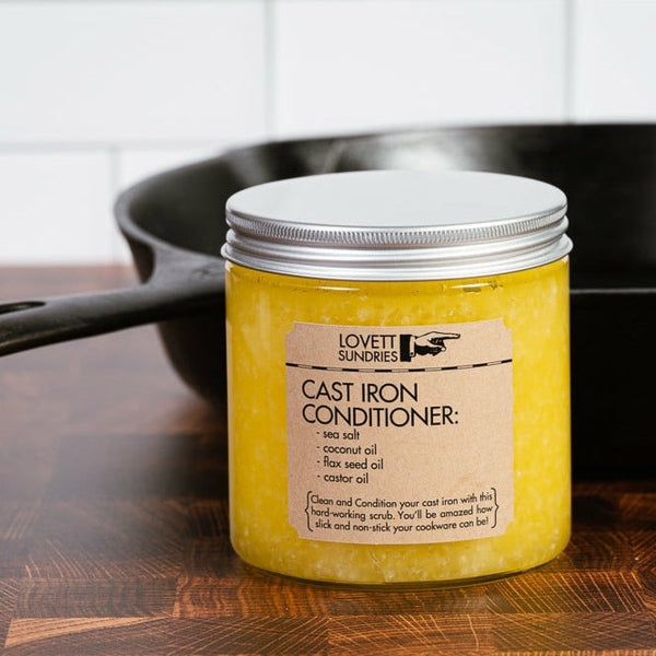 A glass jar with a silver lid holds a yellow substance. In front of a pan, the jar's label reads "Cast Iron Conditioner" and lists "sea salt, coconut oil, flax seed oil, castor oil".