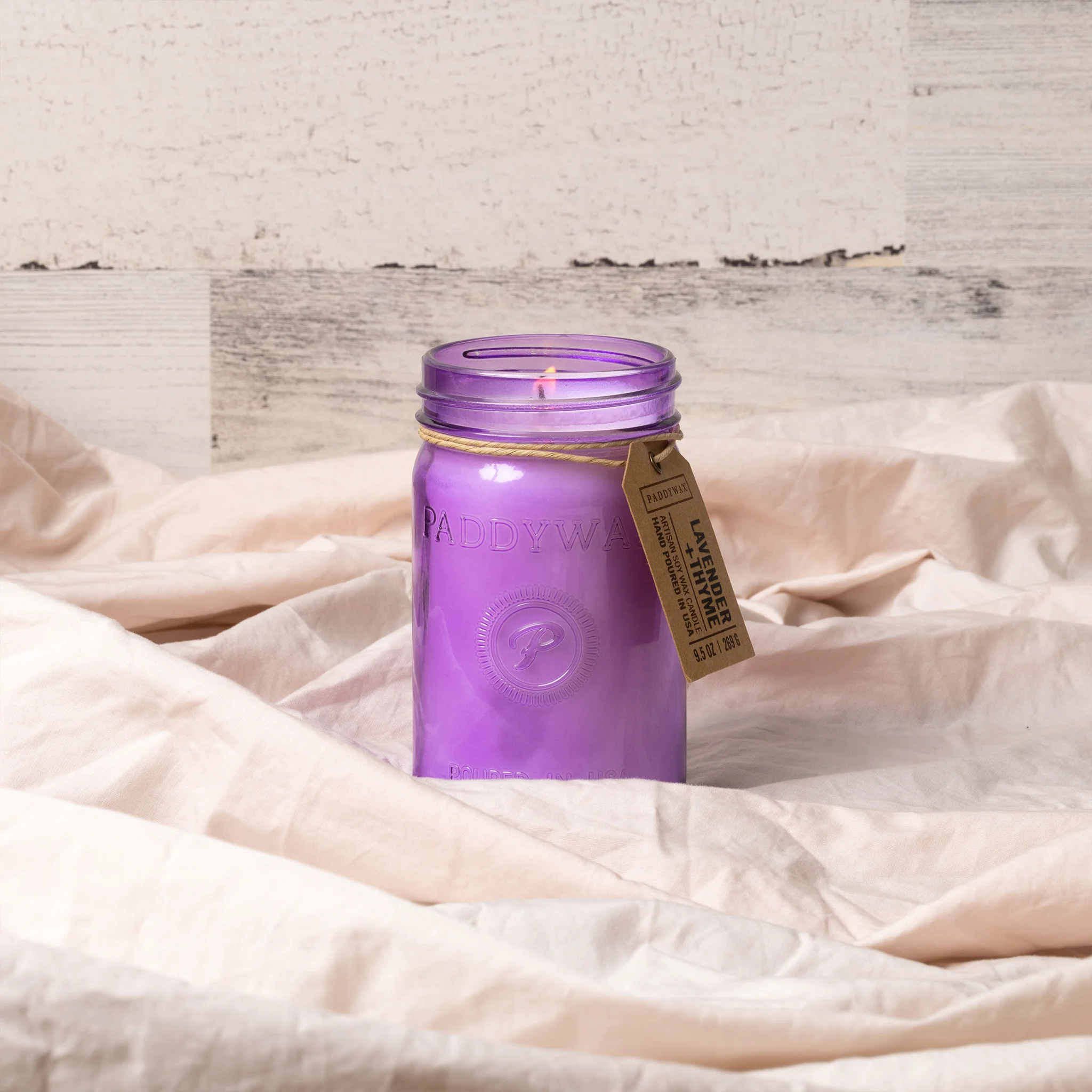 A purple Paddywax artisan soy wax candle in a textured glass jar is shown with the lid off. The label on the lid identifies the scent as "Lavender + Thyme" and notes it's hand-poured in the USA.