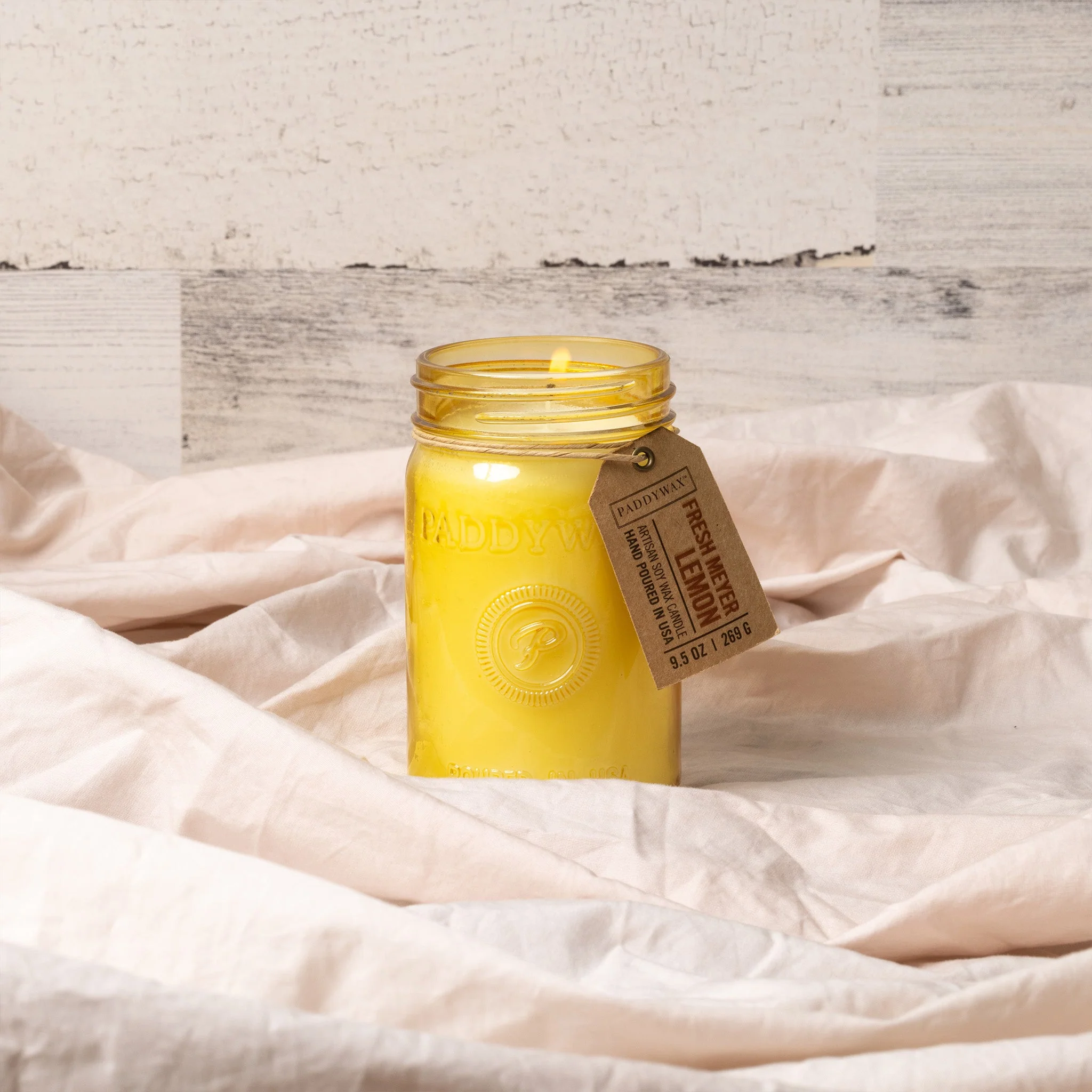 A lit yellow Paddywax "Fresh Meyer Lemon" candle is shown in a jar with its tag attached, resting on tan fabric against a white wooden background.