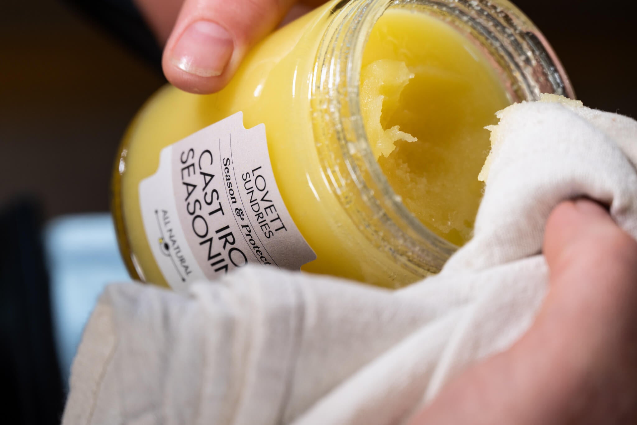 A hand holds a jar of "Lovett Sundries Cast Iron Seasoning" while wiping the side with a white cloth. A scoop has been taken from the yellow contents.
