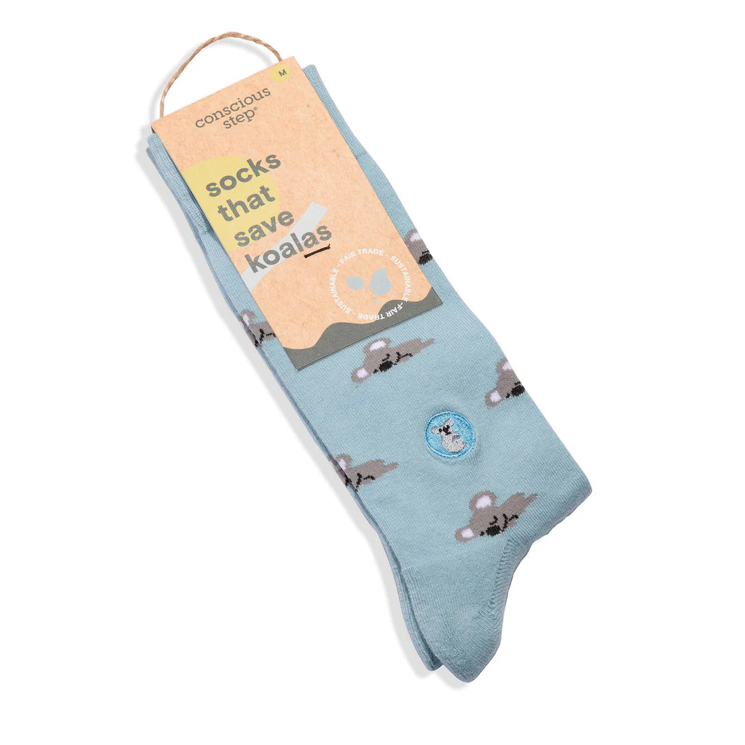 A pair of light blue Conscious Step socks with a pattern of koalas is shown with a tag. The tag identifies them as "socks that save koalas" and notes they are made with fair trade and organic certified cotton.