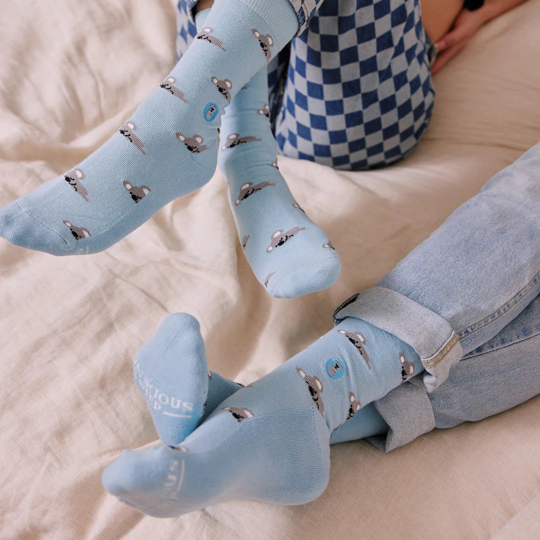 Two people are shown with their legs on a bed. They are both wearing light blue socks with a gray and white koala pattern. The brand name is visible on the bottom of the socks.