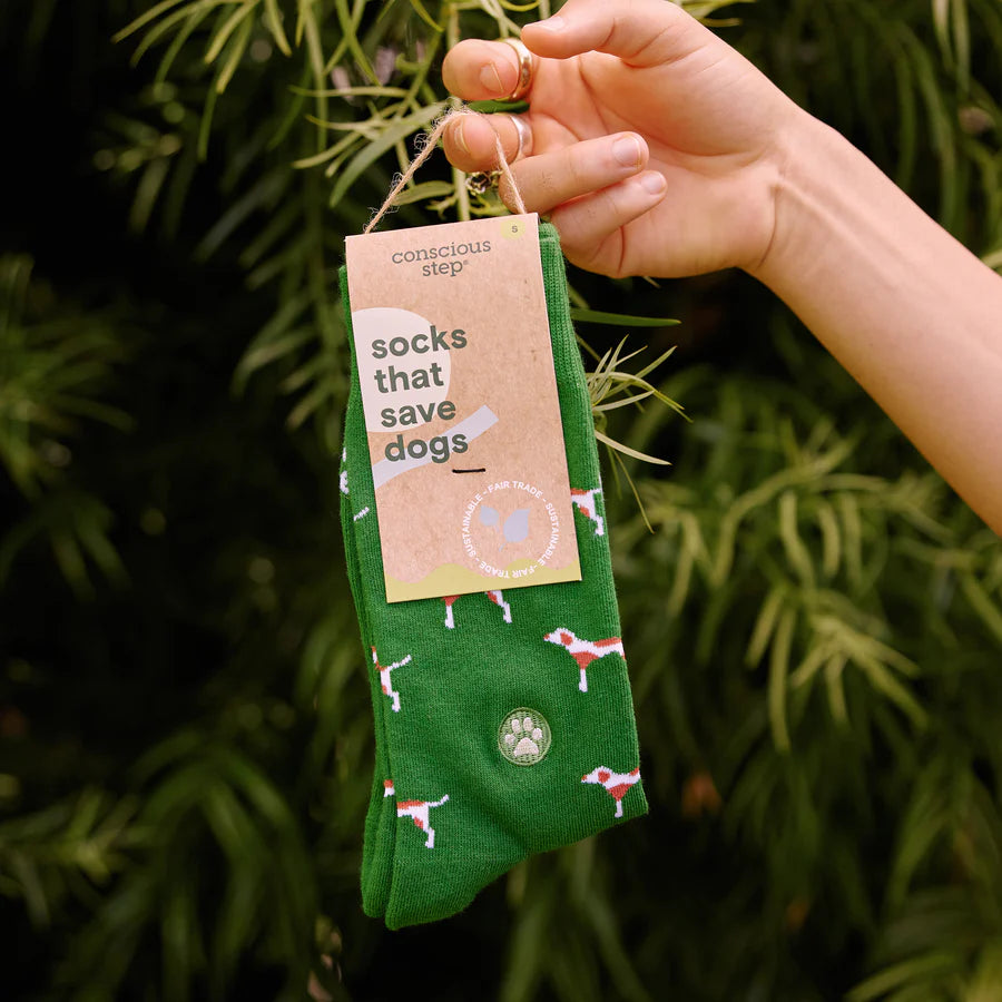 A hand holds a pair of green Conscious Step socks with a red and white dog pattern. The tag on the socks states they are "socks that save dogs" and are made with sustainable, fair trade and organic materials. The background is a blurred green pine foliage.