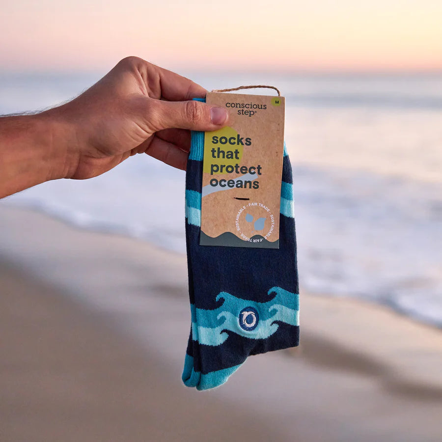 A hand holds a pair of Conscious Step socks with a blue wave pattern against a beach background. The tag identifies them as "socks that protect oceans"