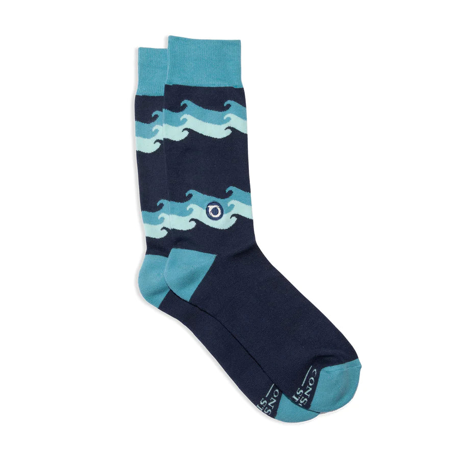 A pair of dark blue Conscious Step socks with a light blue and teal wave pattern is shown against a white background. The socks feature a small circular logo with a wave symbol. The brand name is visible on the bottom of the sock.