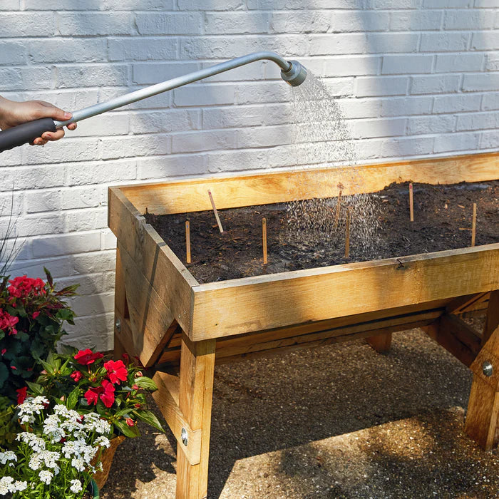 A person's hand waters a raised wooden garden bed on a patio. The water sprays over the freshly planted soil, which has small bamboo sticks to indicate where pops were planted.