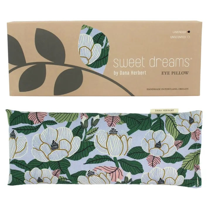 An unscented eye pillow by Dana Herbert with a botanical pattern of white magnolia flowers and green leaves is shown below its cardboard box. The box has a cutout showing the pillow's fabric.